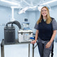 A woman in medical scrubs stands beside a metal cart holding an Arctic Chiller and 13-quart cooling reservoir in a bright operating room. She's attached to the reservoir with the fitted circulating cooling vest. The background shows surgical lights and medical equipment.
