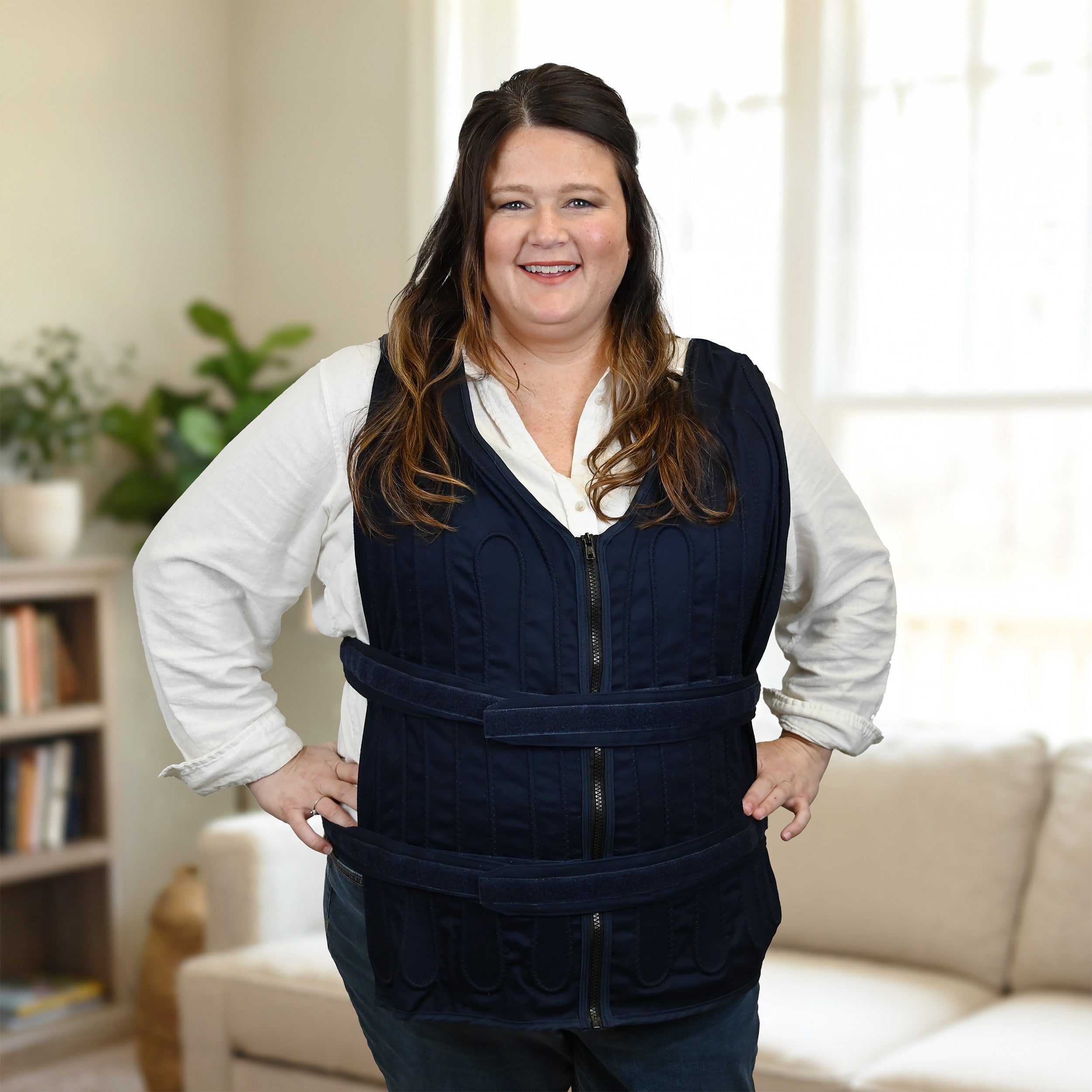 A woman wearing a navy blue adjustable cooling vest over a white collared shirt, standing in a brightly lit living room with a sofa and plant in the background.
