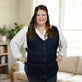 A woman wearing a navy blue adjustable cooling vest over a white collared shirt, standing in a brightly lit living room with a sofa and plant in the background.