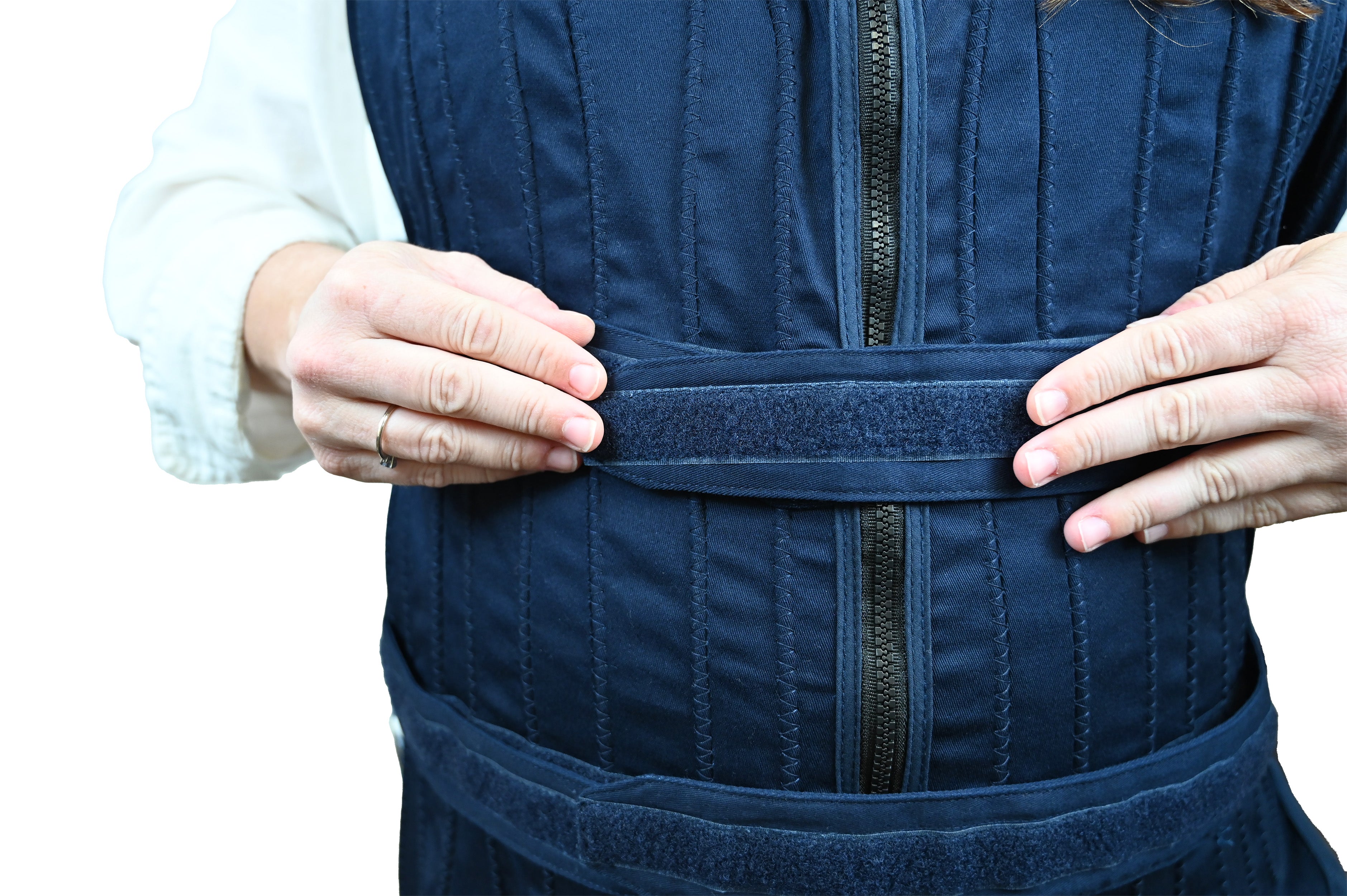 A close-up of a person wearing a dark navy blue circulating cooling vest, with both hands adjusting a horizontal strap across the torso. The garment has vertical stitched panels and a front zipper, shown against a plain white background.
