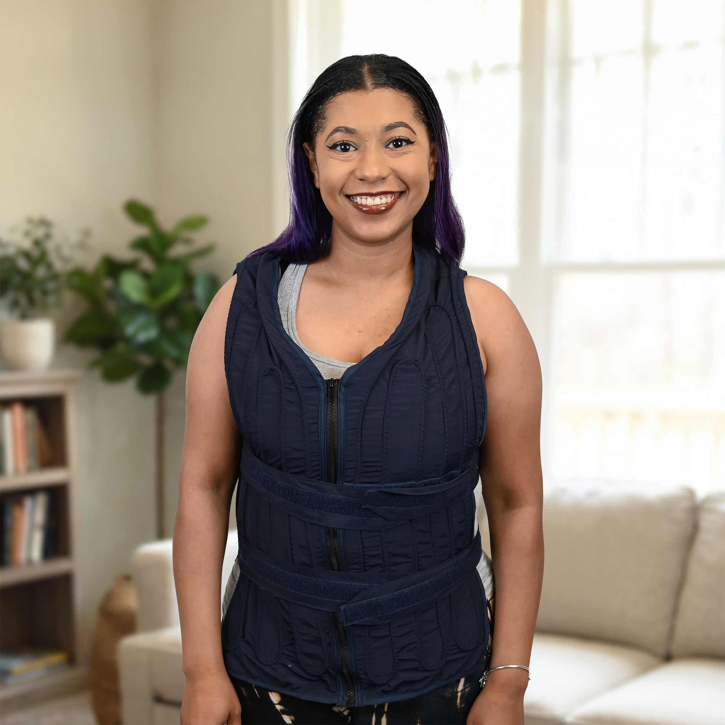 A smiling woman with purple-streaked black hair stands in a living room wearing a dark navy adjustable circulating cooling vest over a gray tank top. The room has a light-colored sofa, a bookshelf, and a potted plant in the background.