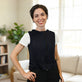 A smiling woman with short dark hair stands indoors wearing a fitted circulating cooling vest over a white short-sleeve top and black pants. The background shows a softly lit living room with a sofa, window, and plant.