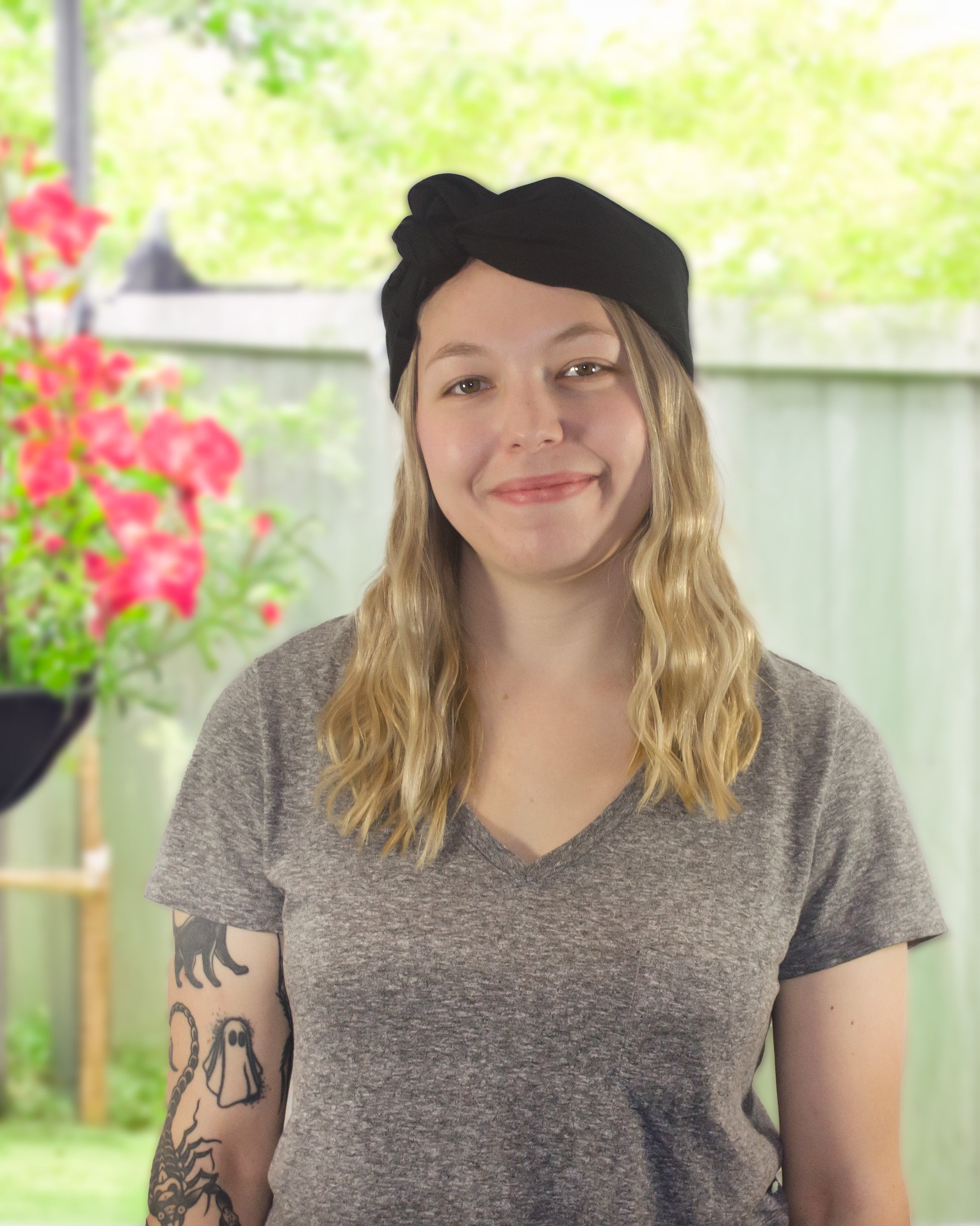 Person wearing a black cap and gray t-shirt with tattoos, standing outdoors with flowers and greenery in the background.