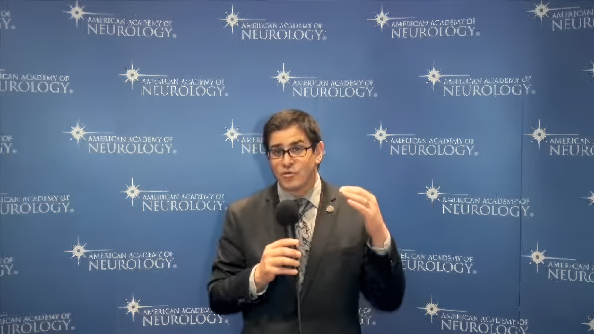 Man speaking into a microphone in front of an American Academy of Neurology backdrop