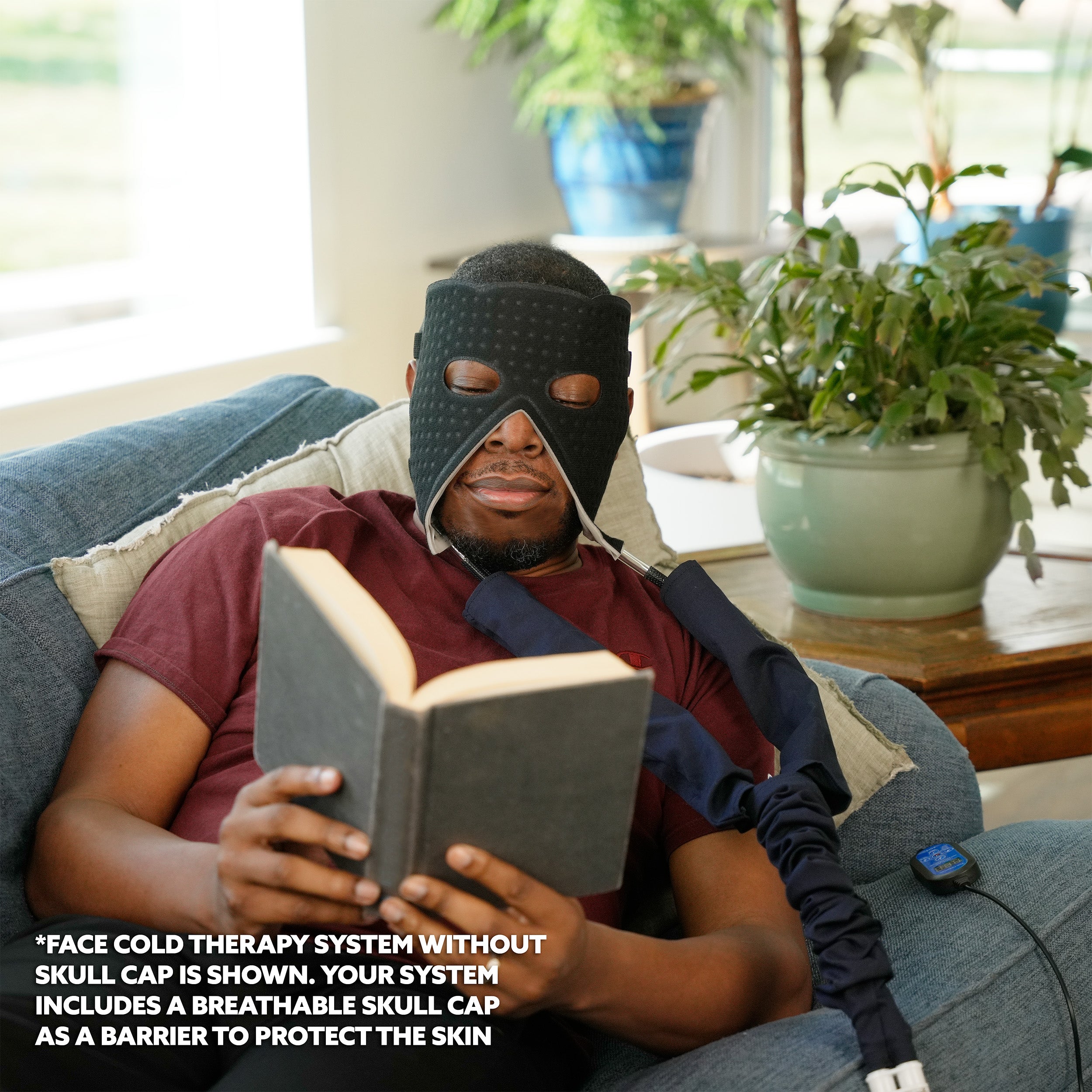 Person wearing a face cold therapy pad and reading a book in a home setting with plants and furniture.
