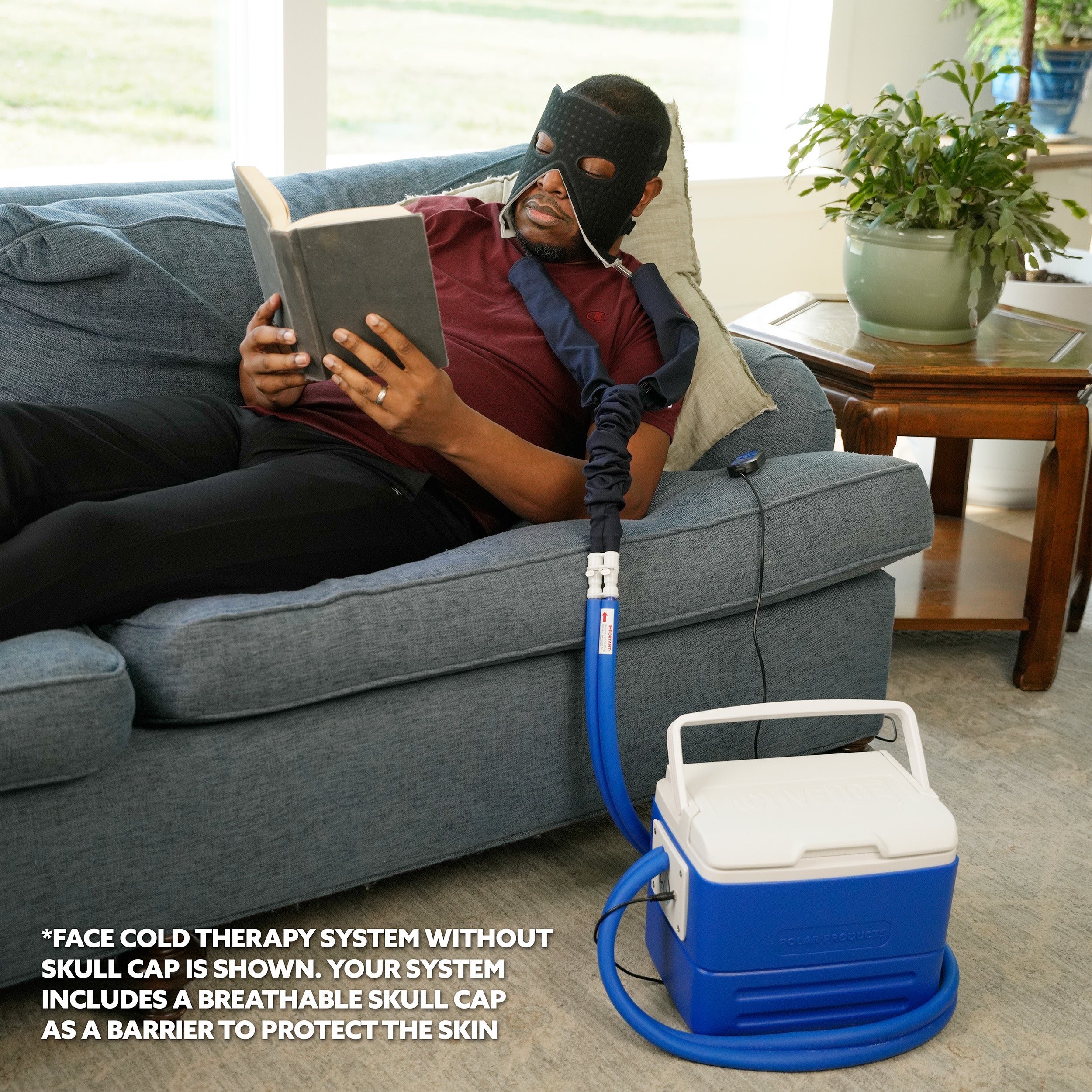 Person reading a book with a face cold therapy system attached, sitting on a couch in a living room.