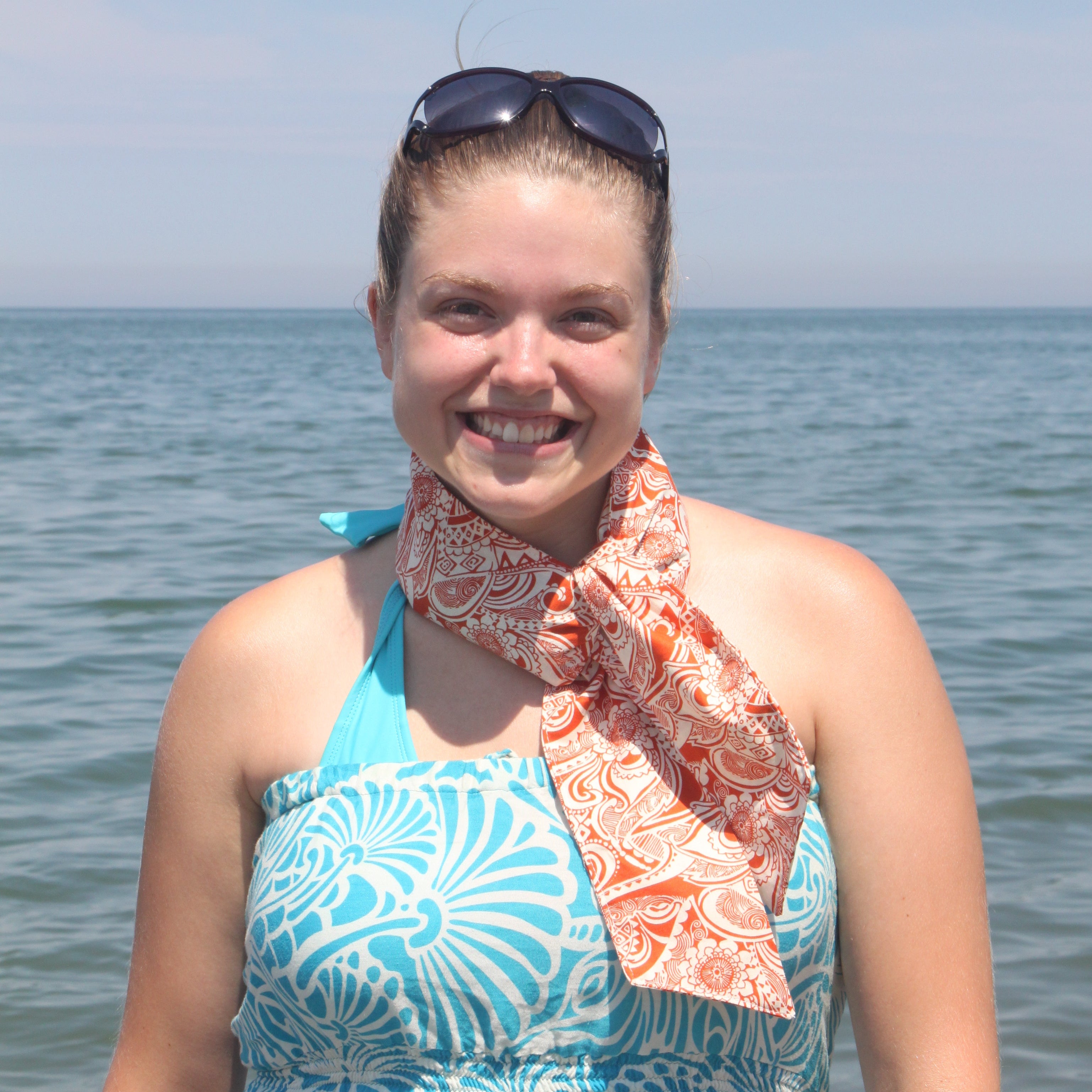 Woman in a patterned dress standing by water with orange Fashion Cooling Scarf around her neck. 