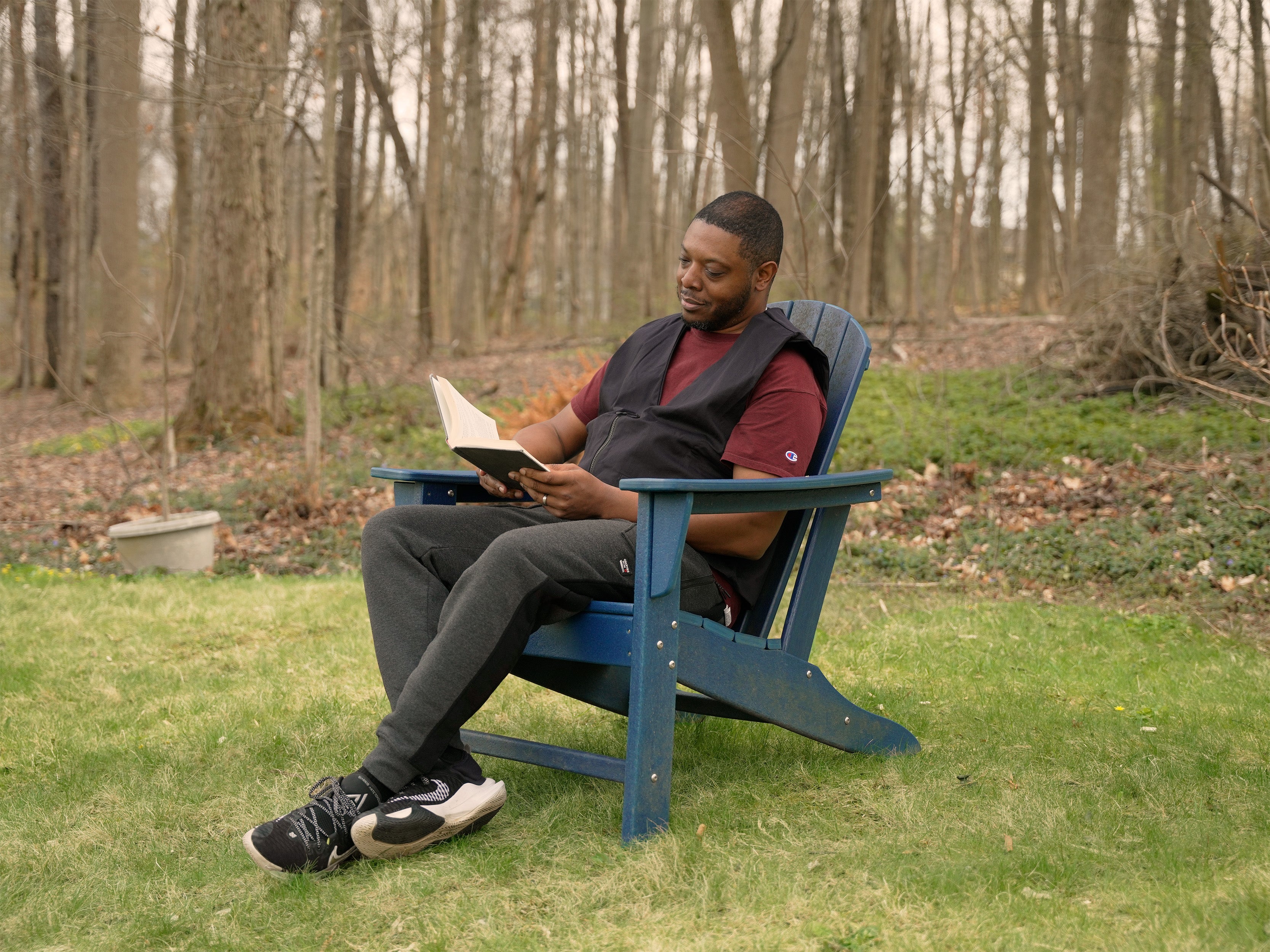 Man sitting in a blue Adirondack chair reading a book in a forest setting wearing a black fashion cooling vest.