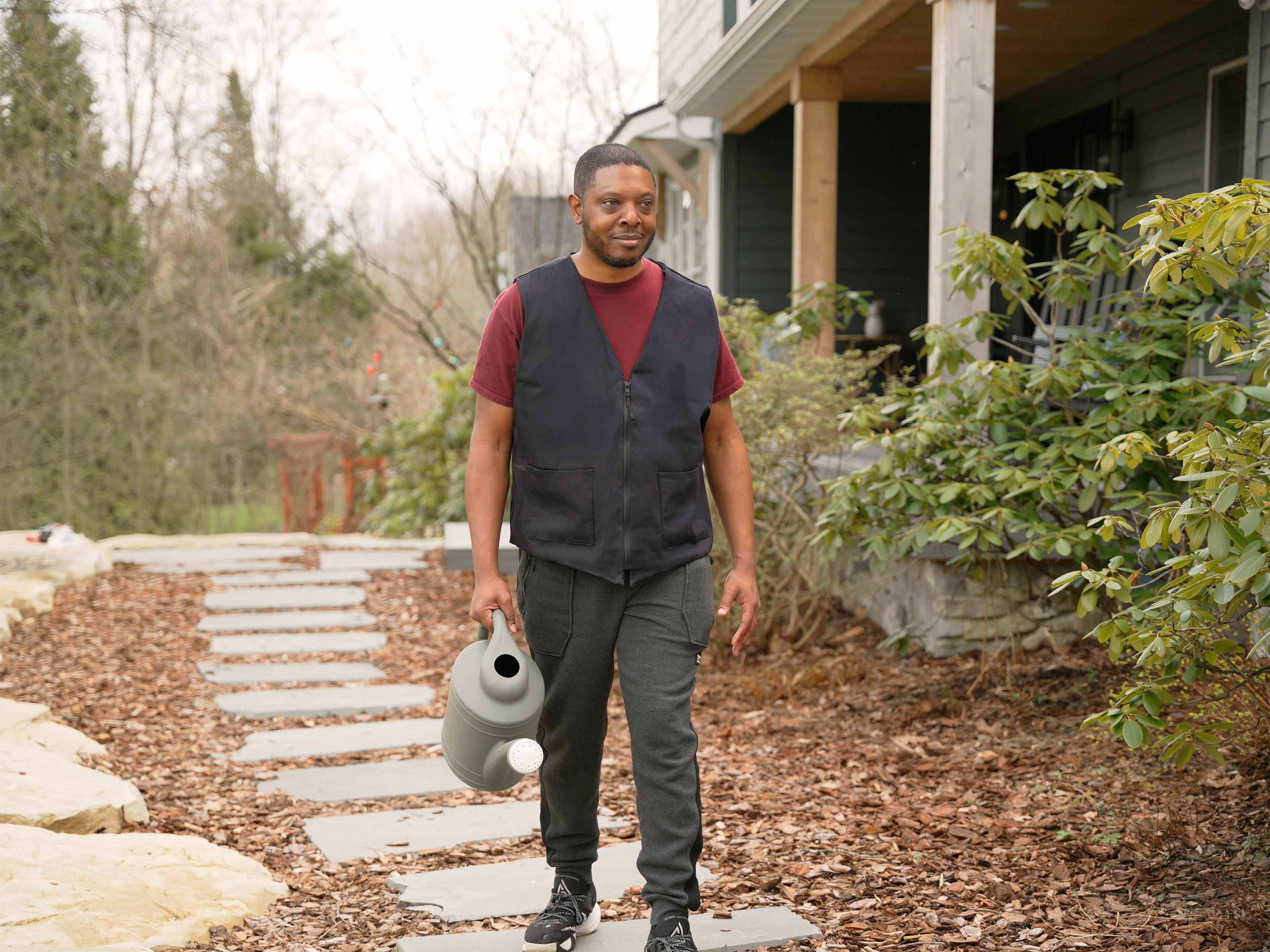 Man holding a watering can on a stone path in front of a house wearing a black fashion cooling vest