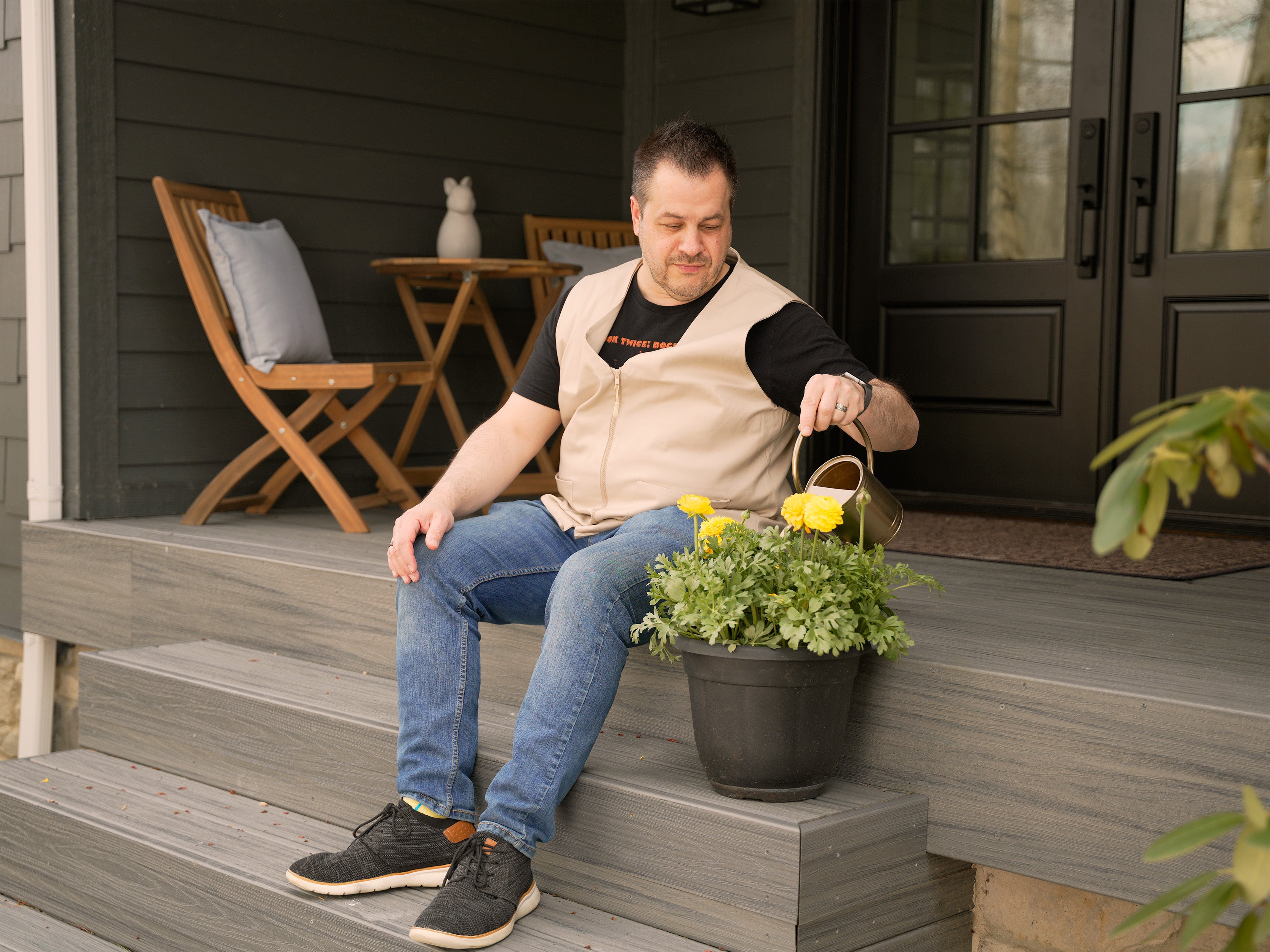 Man sitting on a porch watering a potted plant while wearing a beige fashion cooling vest.