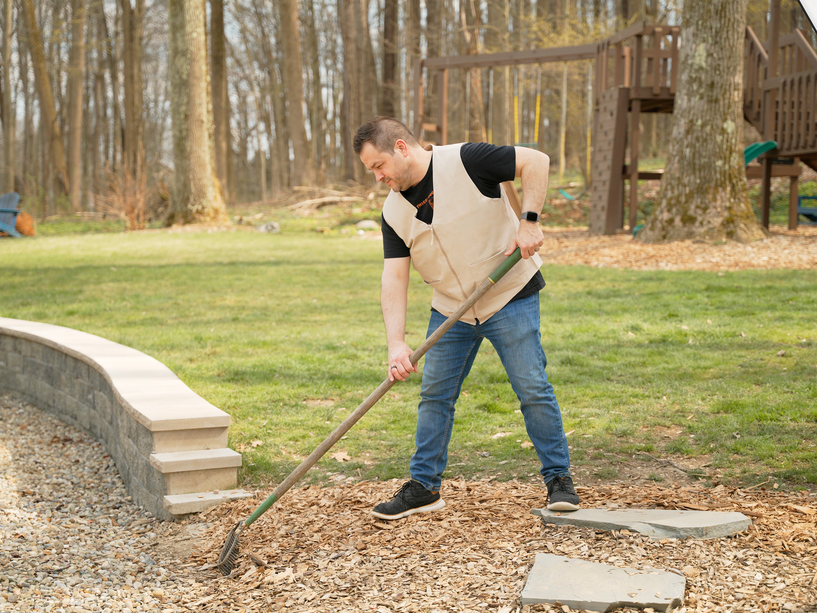 A man wearing a beige adjustable cooling vest over a black t-shirt and blue jeans is raking wood chips in an outdoor backyard setting with trees and a wooden playset in the background.