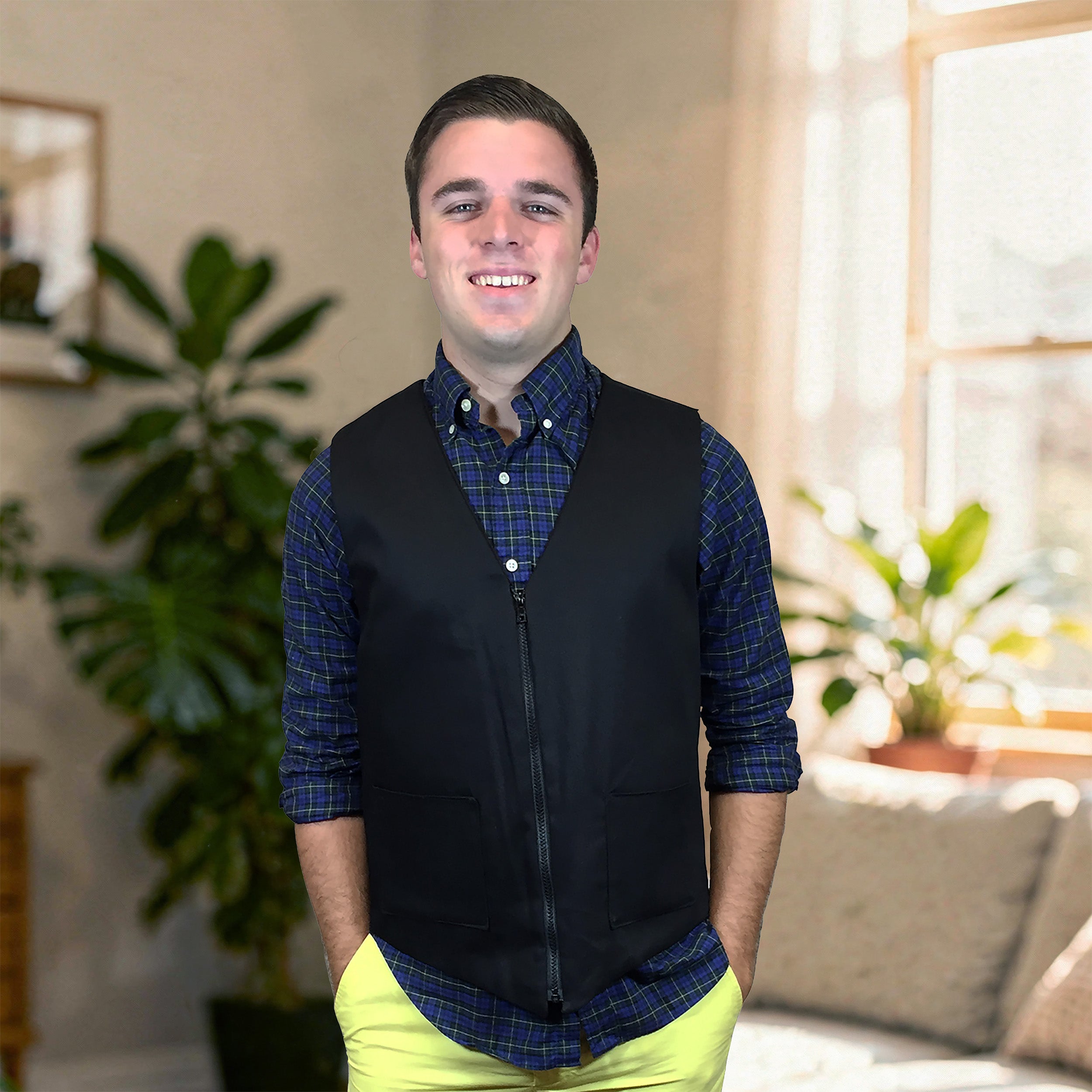 A smiling young man standing indoors wearing a black fashion cooling zip-front vest over a blue and black plaid button-down shirt, with bright yellow pants. The background shows a softly blurred living room with plants, a window, and a sofa.