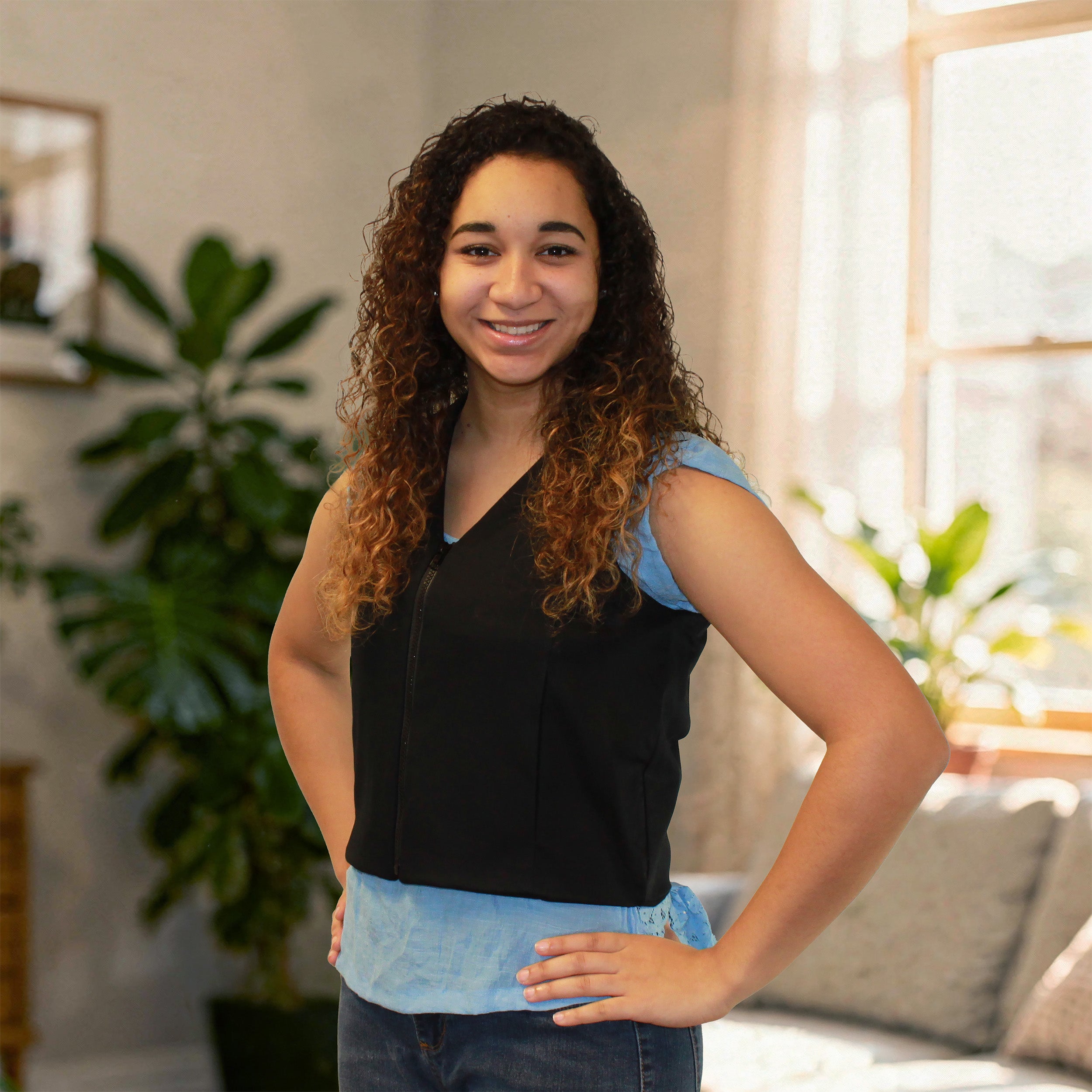 A smiling woman with curly brown hair stands indoors wearing a sleeveless black fashion cooling vest over a light blue top and dark jeans. The photo is taken in a softly lit room with plants and a window in the background.