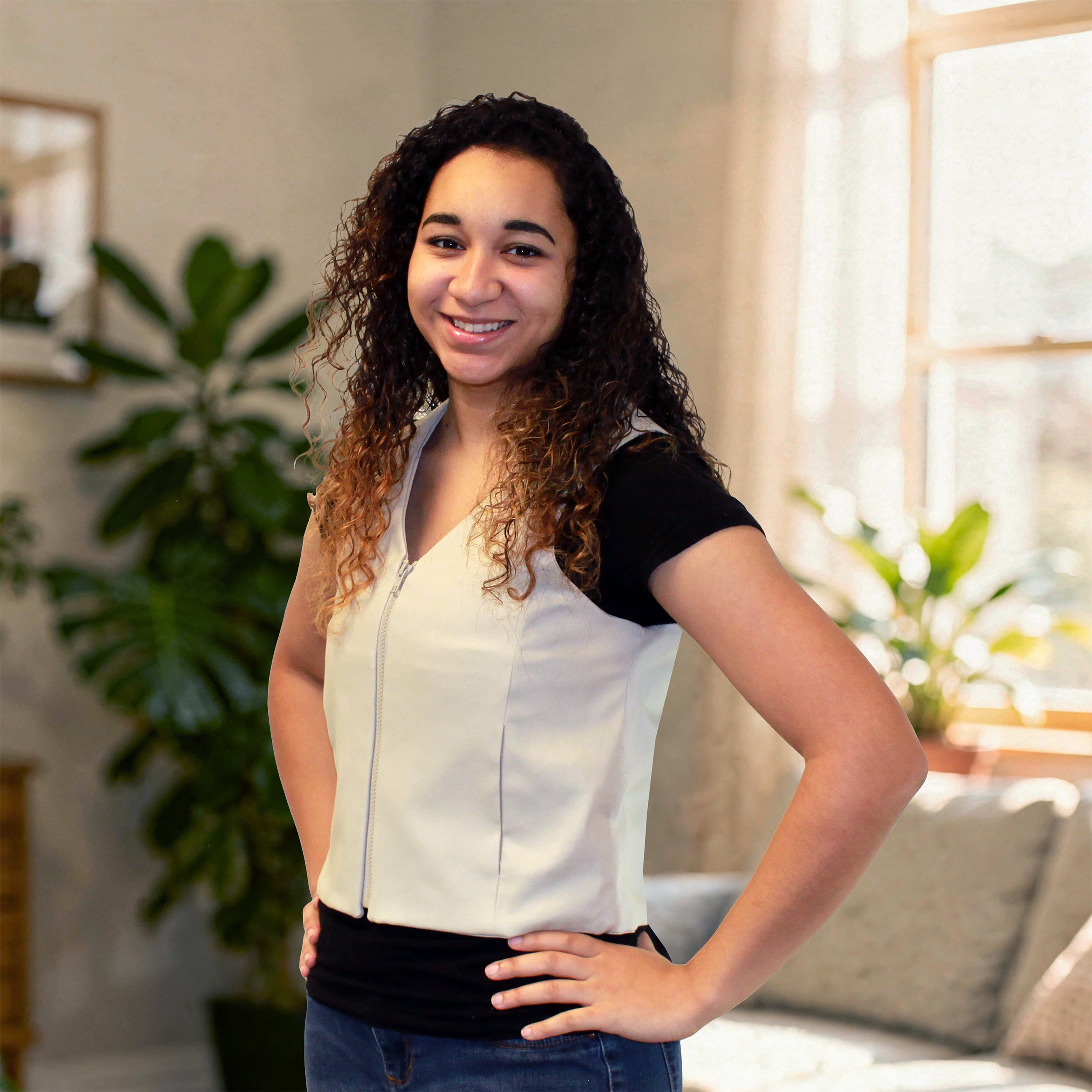 A smiling woman with curly brown hair stands indoors wearing a sleeveless khaki fashion cooling vest with a black short-sleeve layer underneath, paired with dark blue jeans. The background shows a softly lit room with plants, a window, and a sofa.