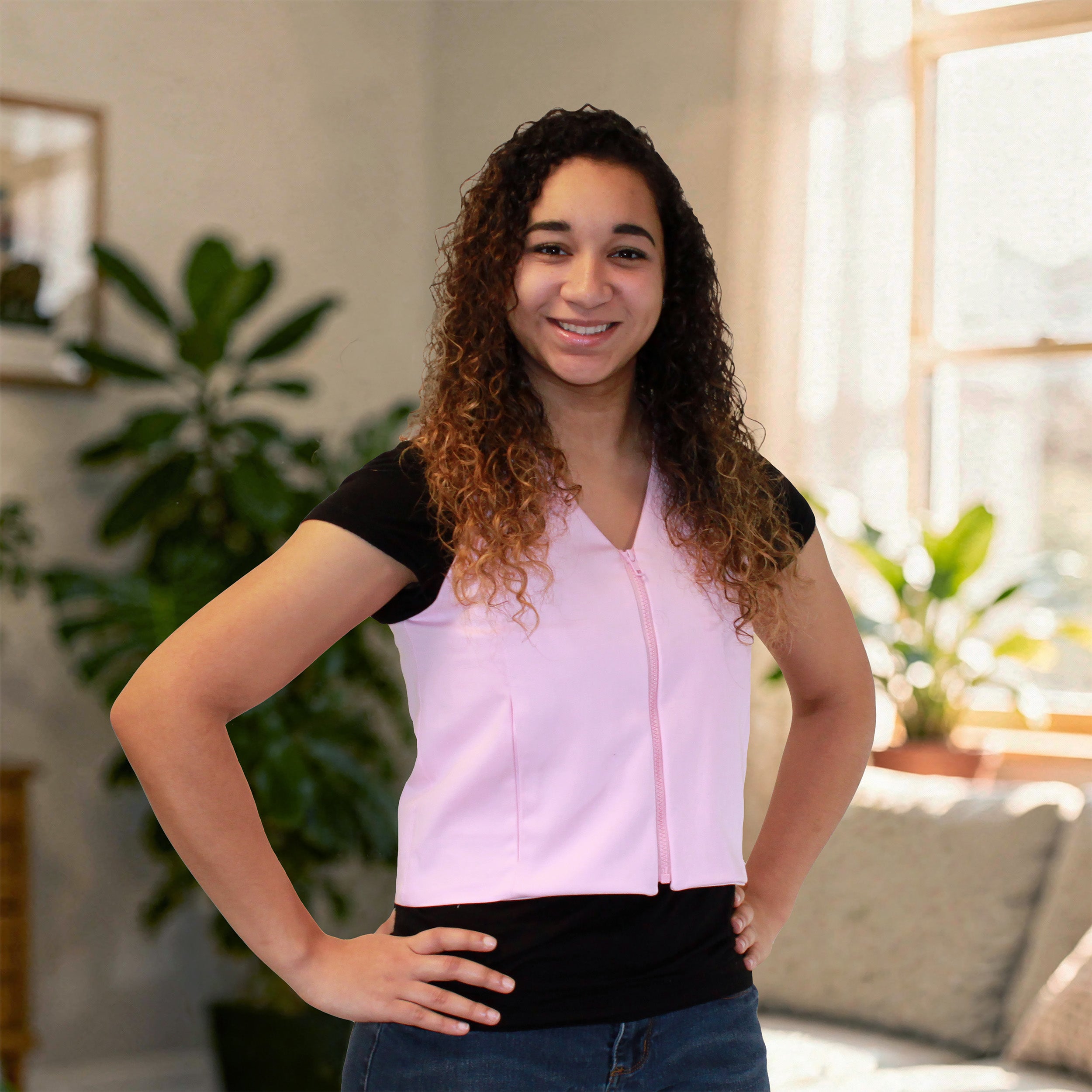 A woman with curly brown hair stands indoors wearing a light pink sleeveless fashion cooling zip-front vest over a black short-sleeve top and blue jeans. The photo is taken in a softly lit room with plants and a window in the background.