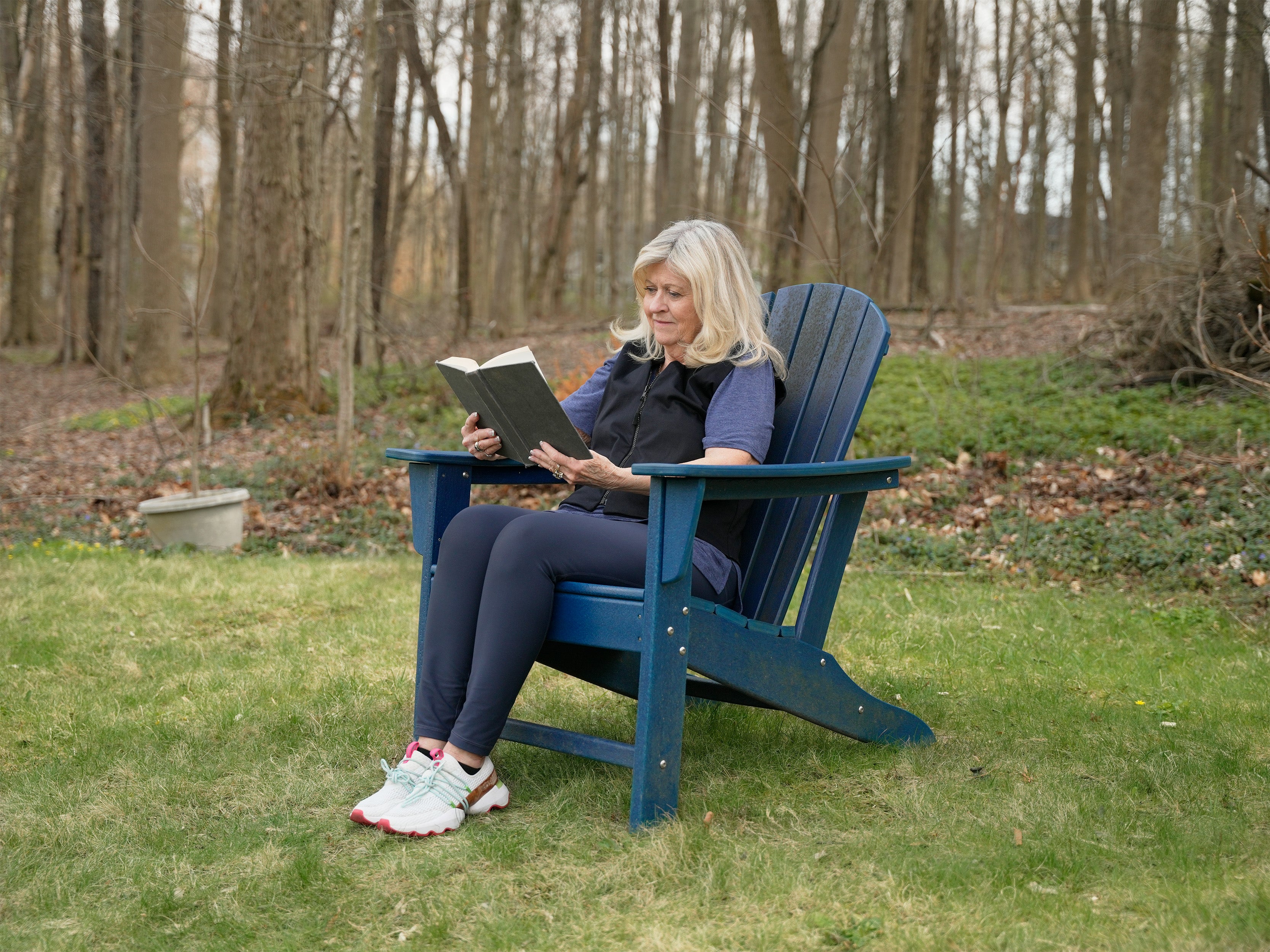 Woman reading a book in a blue Adirondack chair outdoors in a wooded area wearing a black fashion cooling vest.