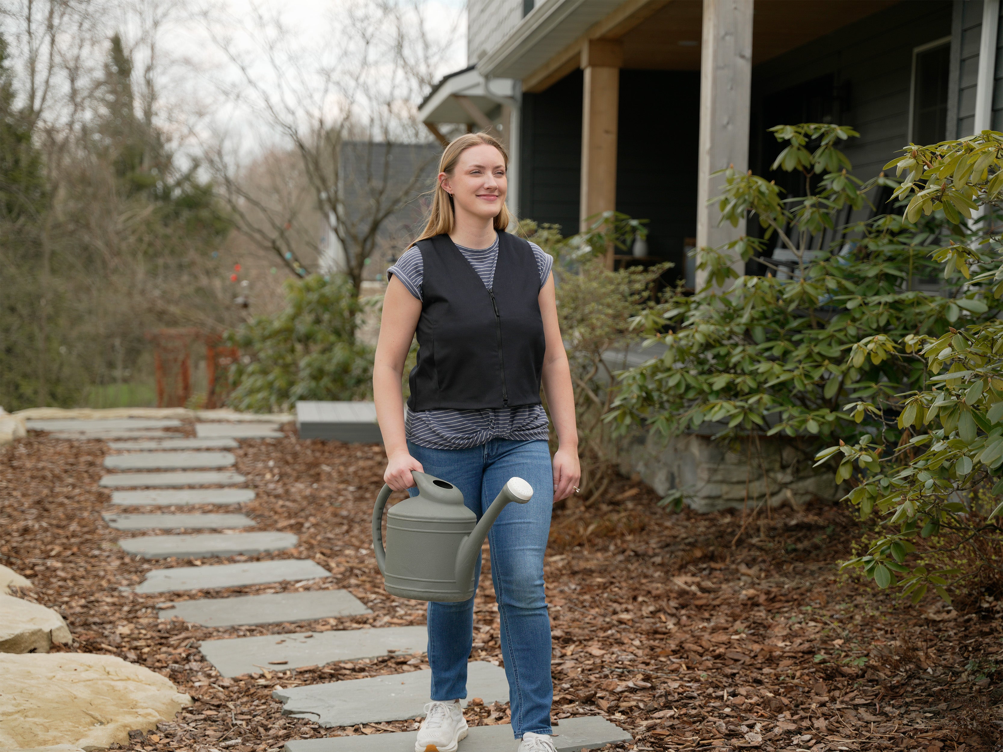 A woman walking on a stone path in front of a house, holding a gray watering can wearing a black fashion cooling vest.