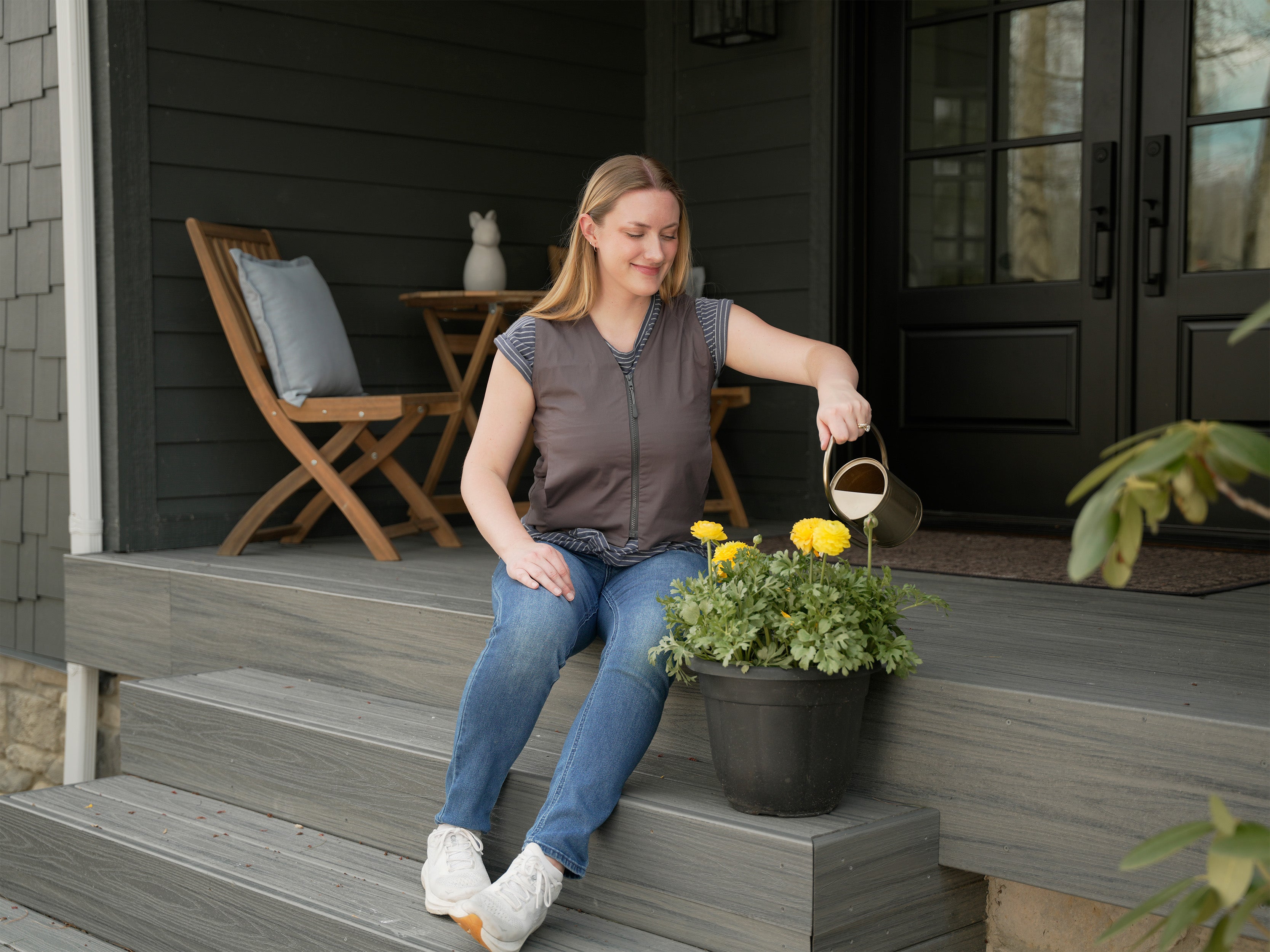 Woman sitting on a porch with a potted plant and watering can wearing a gray fashion cooling vest