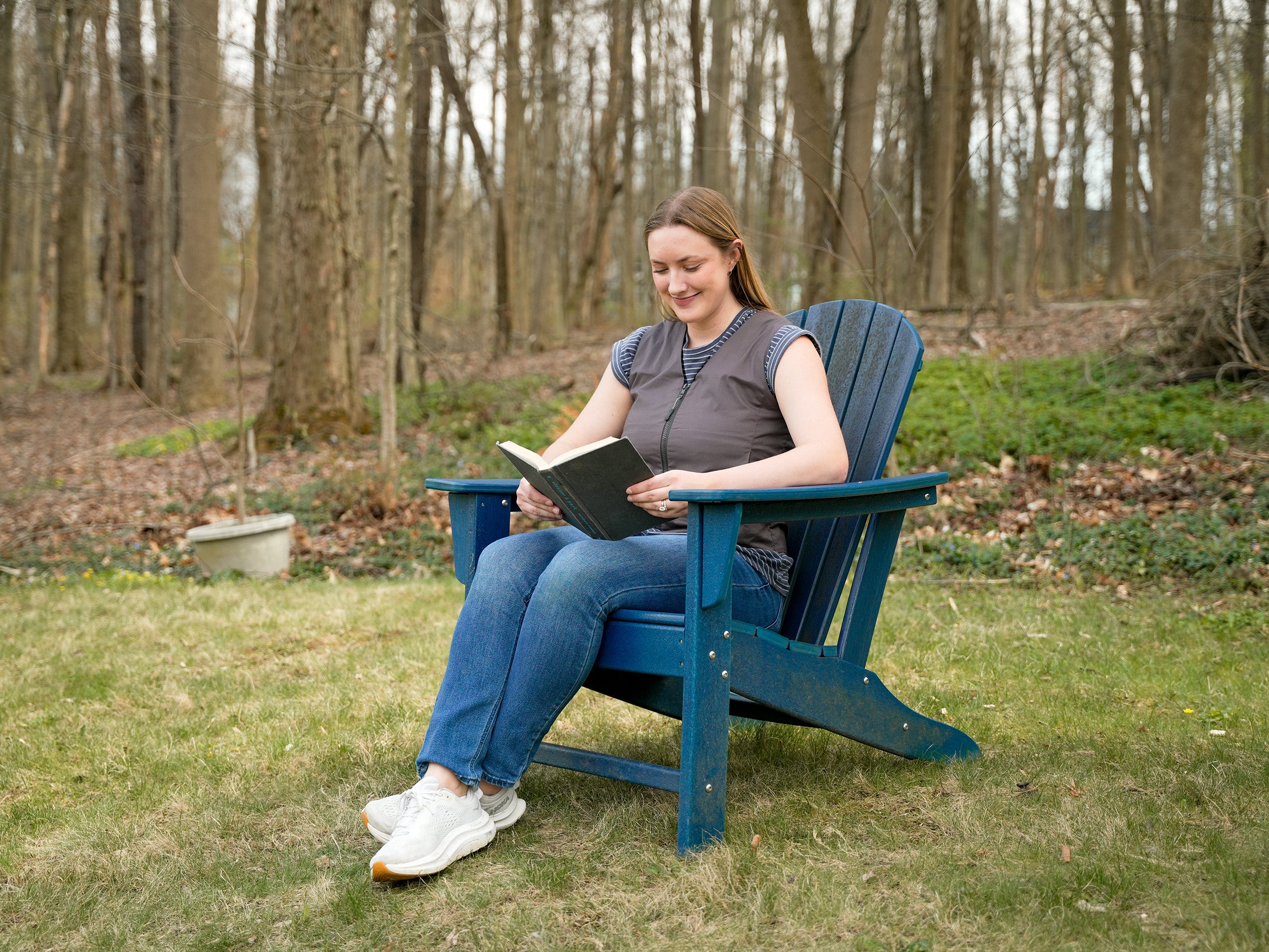 A woman sits in a blue Adirondack chair on a grassy lawn, reading a black book in an outdoor wooded setting wearing a gray Fashion Cooling Vest.