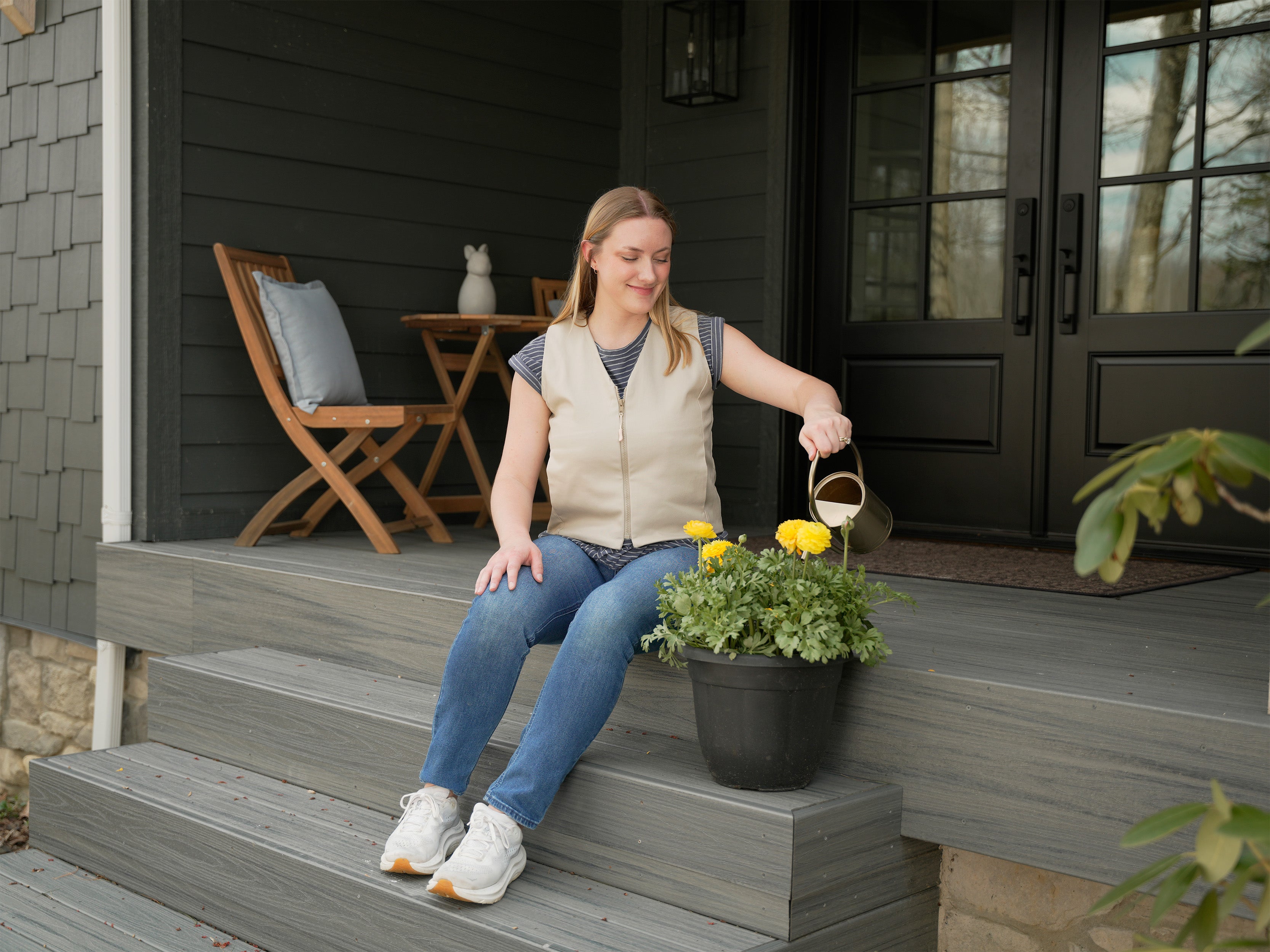 Woman sitting on a porch steps with a potted plant, holding a small watering can wearing a khaki fashion cooling vest