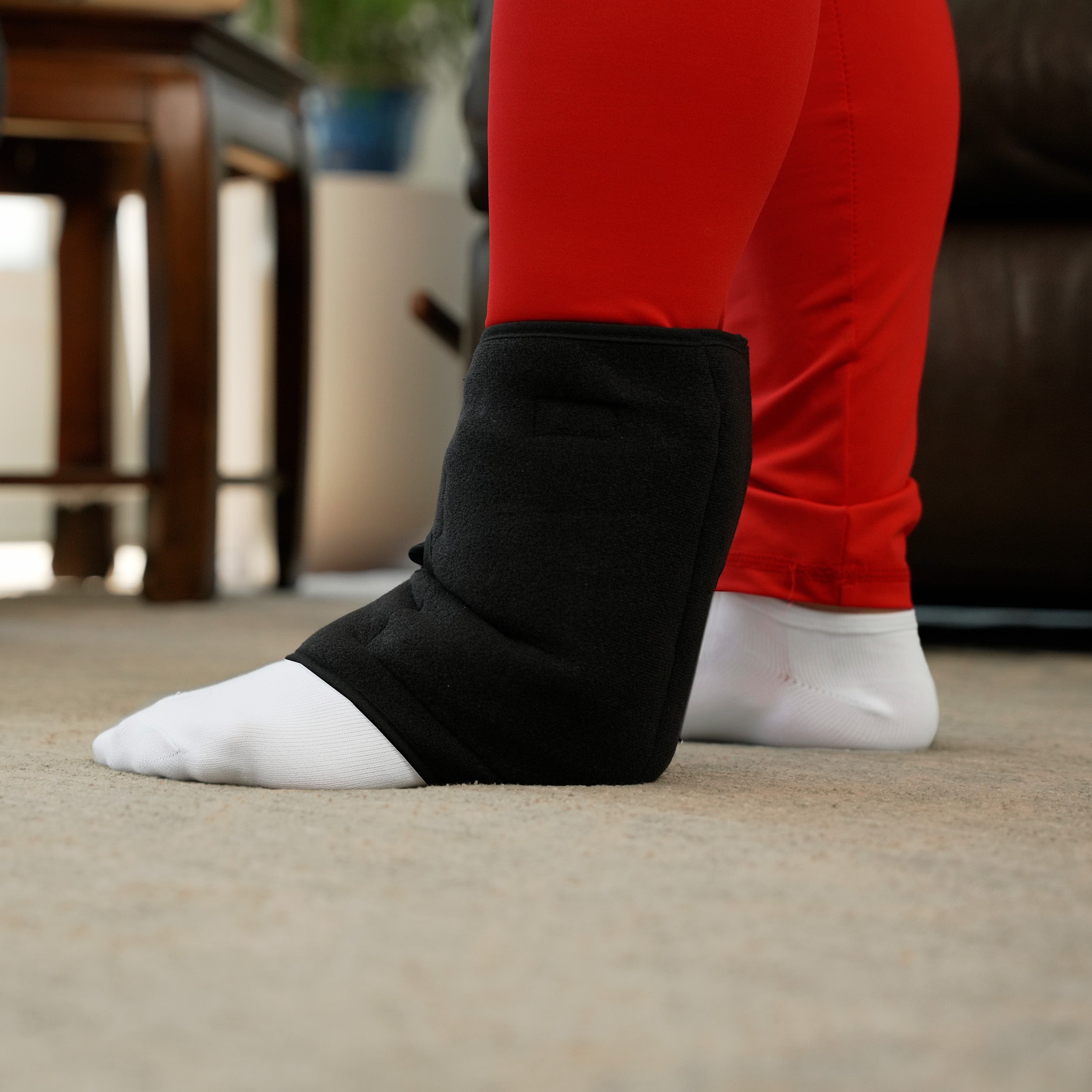 Person wearing black Soft Ice® Ankle Wrap over white socks in a home setting.