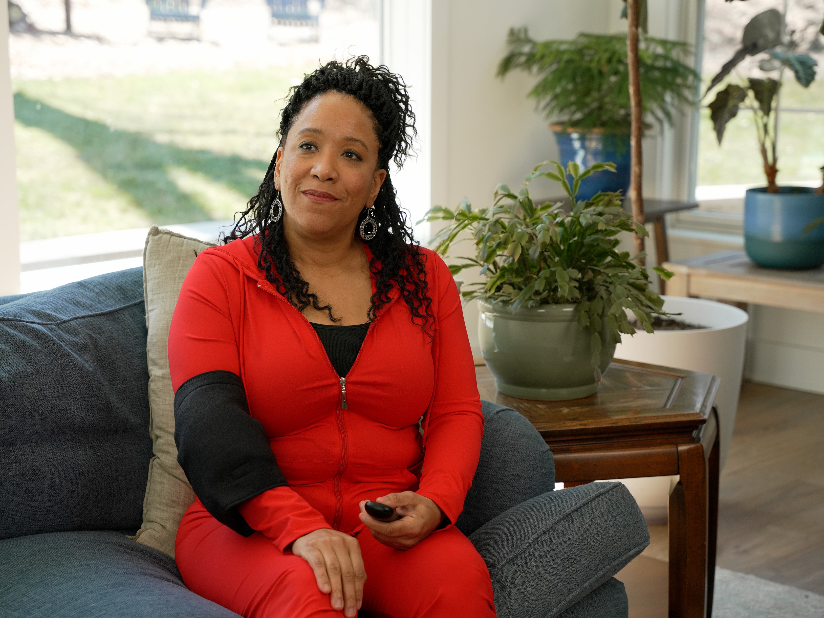 Woman in a red outfit sitting on a couch in a living room with a Soft Ice® Elbow Wrap on with plants and a window in the background.