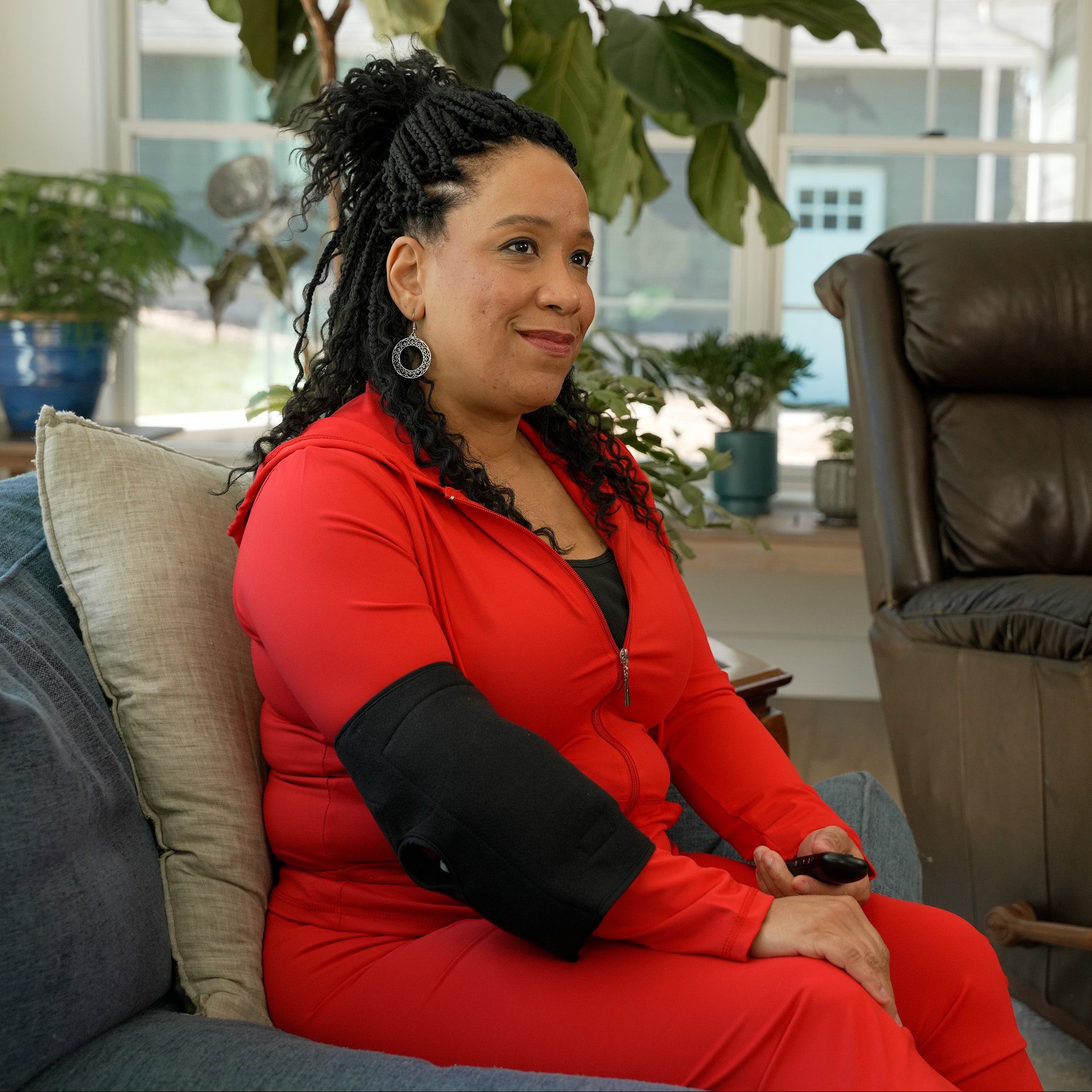 Woman in red outfit sitting on a couch with a Soft Ice® Elbow Wrap on with a brown leather recliner chair in the background.