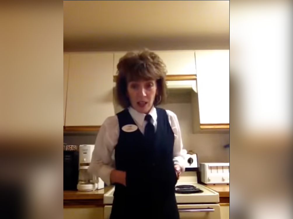 A woman with short brown hair stands in a kitchen wearing a white collared shirt, a black fashion cooling vest, and a black tie. She appears to be speaking or presenting, with kitchen cabinets, a stove, and countertop items visible in the background.