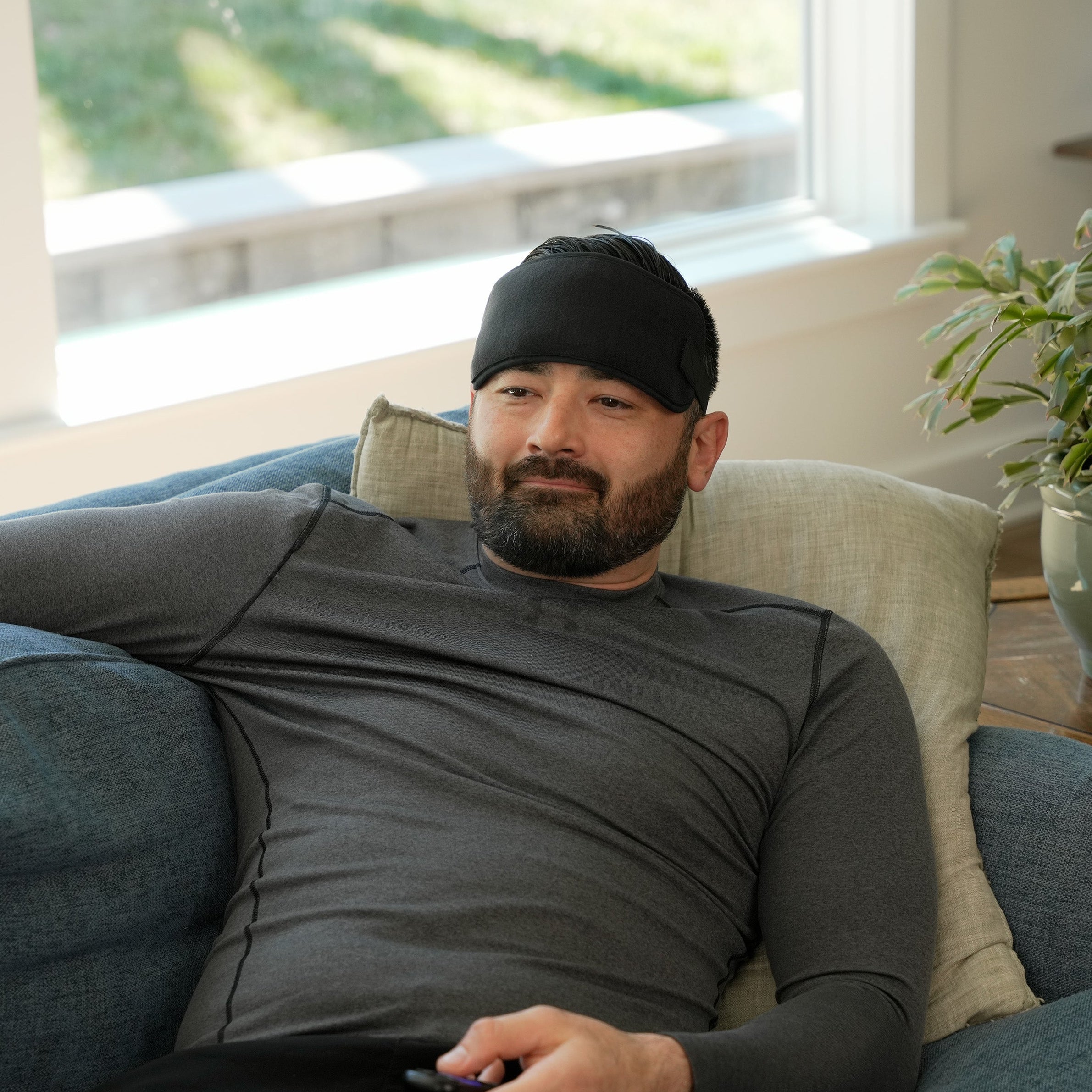 Man sitting on a couch wearing a black Soft Ice® Head and Jaw Wrap and gray shirt, holding a remote control.