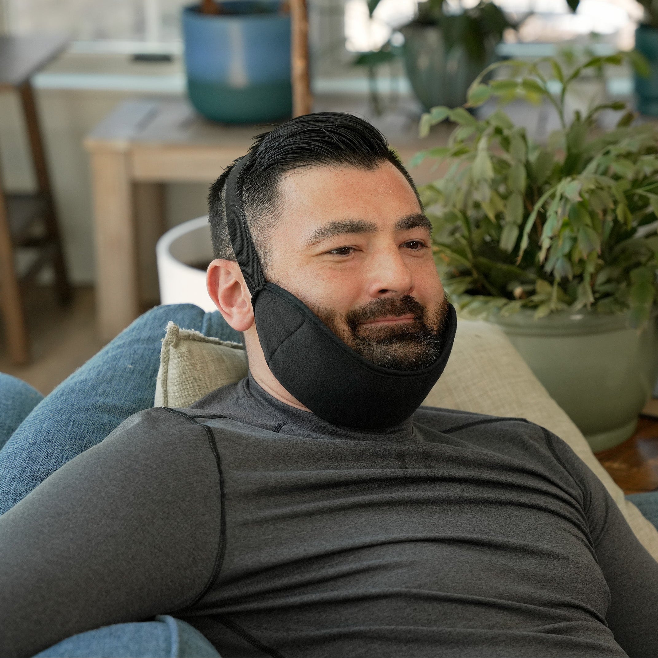 Man wearing a Soft Ice® Jaw Wrap in a living room with plants and furniture.