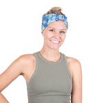 A smiling woman with blonde hair tied up in a blue and white patterned Cooling headband, wearing a gray ribbed tank top against a solid white background.