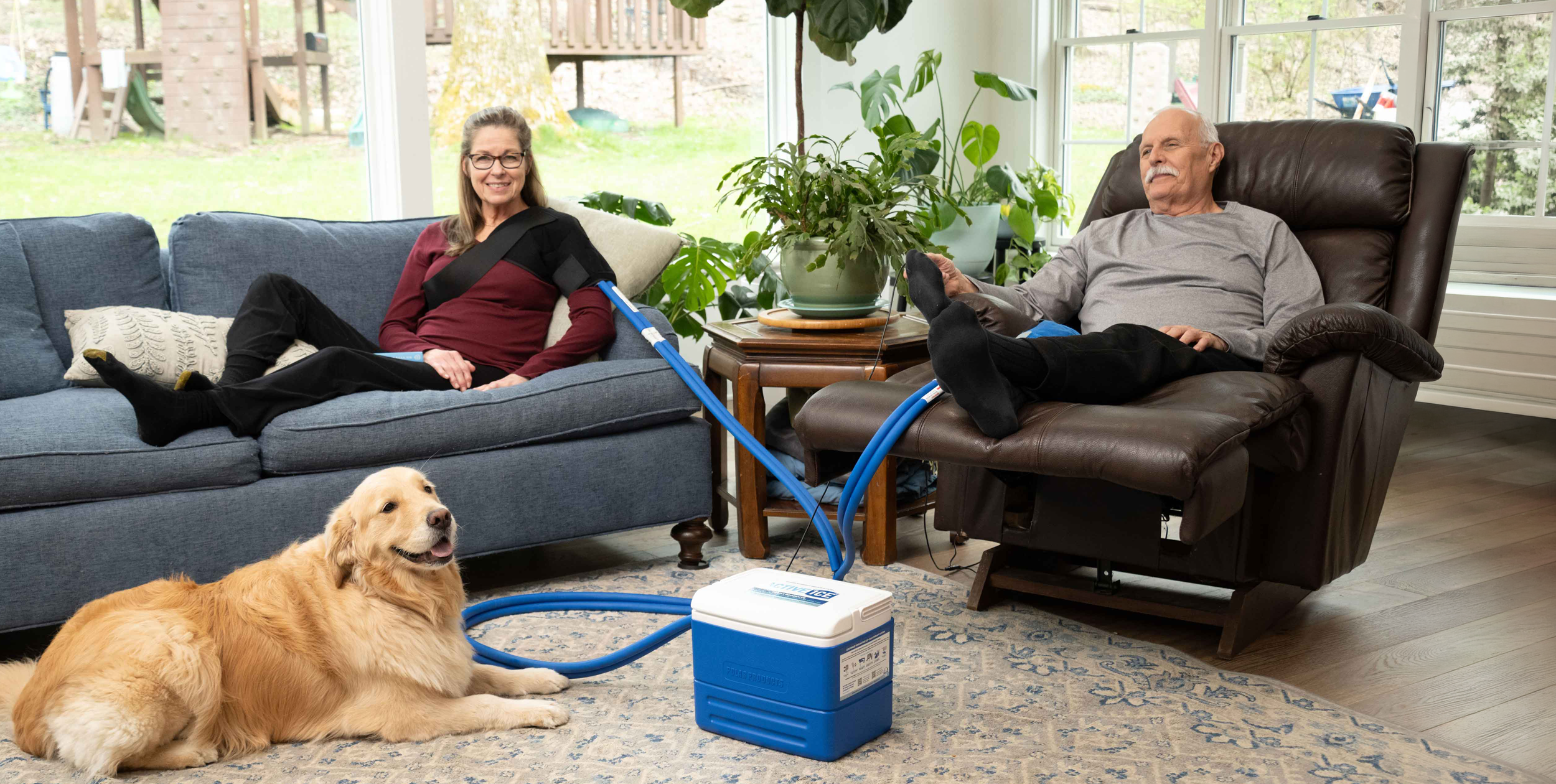 A man relaxes in a brown recliner while a woman sits on a blue sofa, with a golden retriever dog lying on the floor nearby. A blue and white portable cooling unit with connected tubing is placed on the rug in the living room.