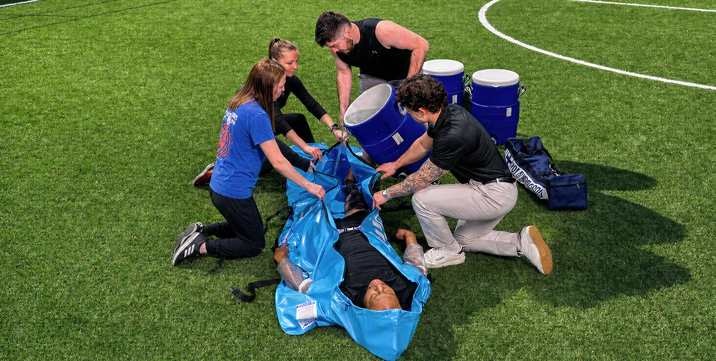Several people on a green turf field are gathered around a person lying on a bright blue Polar Life Pod®, with blue water barrels and bags nearby.