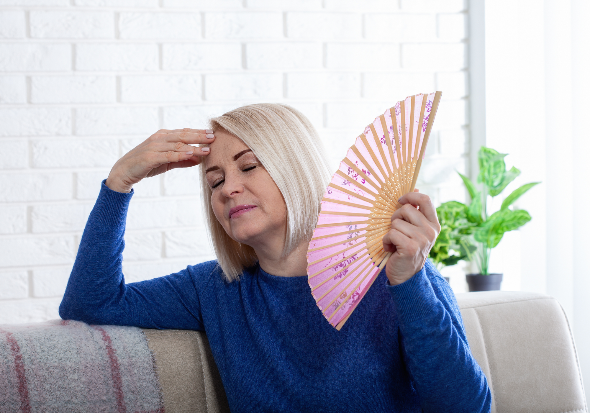 Woman in blue sweater holding a pink folding fan, sitting on a couch with a white brick wall and plant in the background.