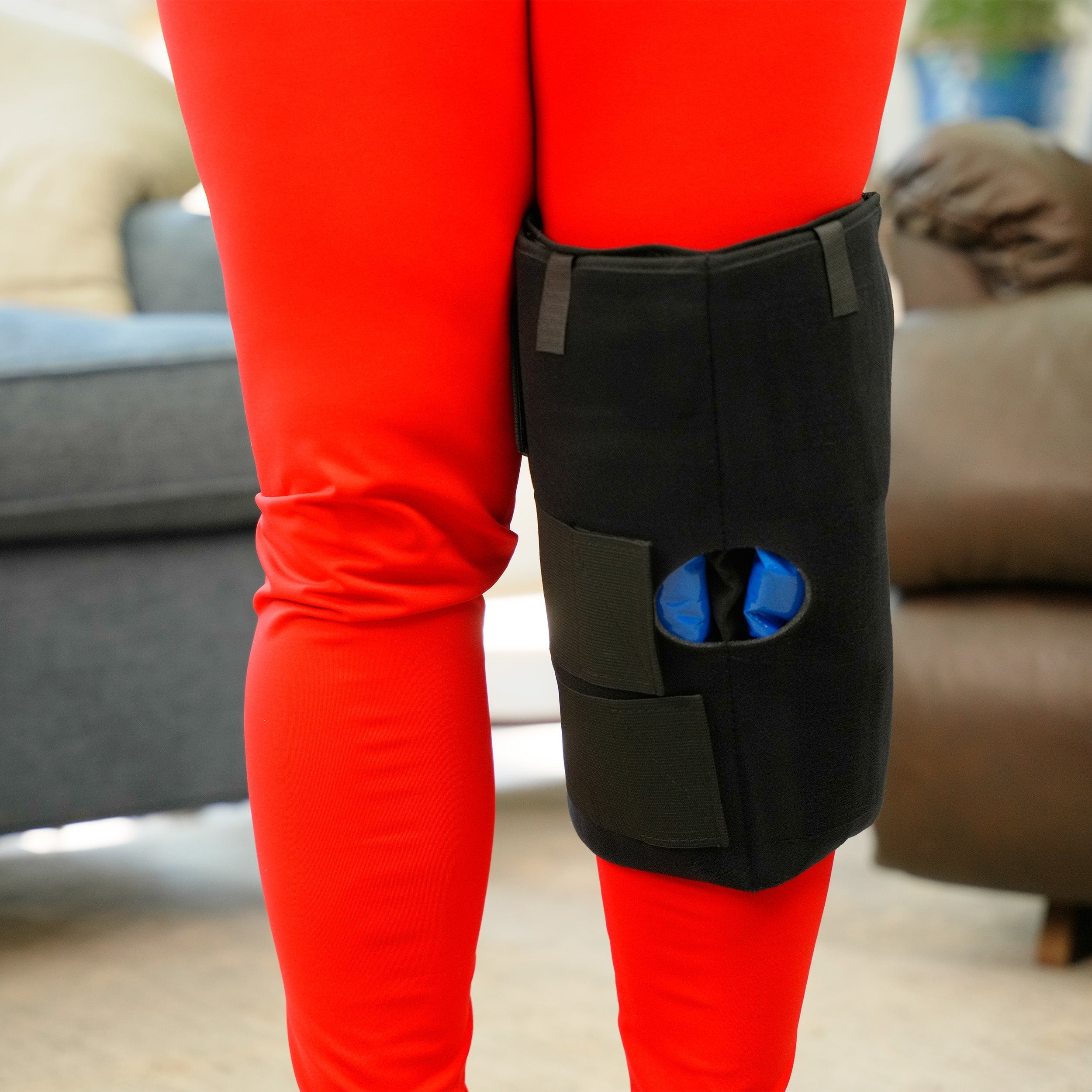 Person wearing a black Soft Ice® Knee Wrap with blue packs on red pants in a living room setting.
