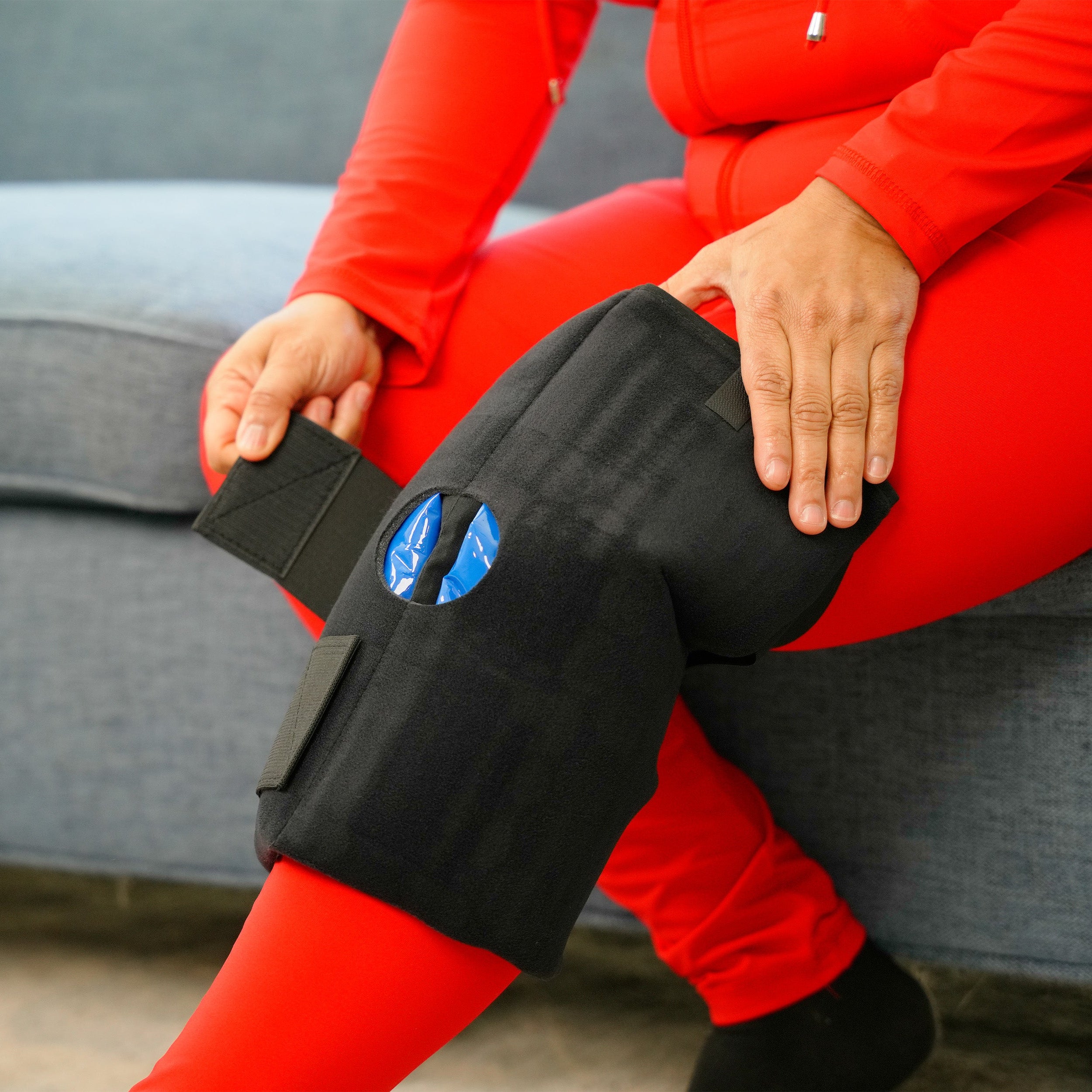 Woman in red outfit applying a Soft Ice® Knee Wrap on a gray couch.