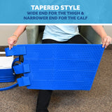 A person seated in an armchair holds a bright blue tapered full circumference knee cold therapy pad, with text at the top reading 'TAPERED STYLE' and 'WIDE END FOR THE THIGH & NARROWER END FOR THE CALF.'.