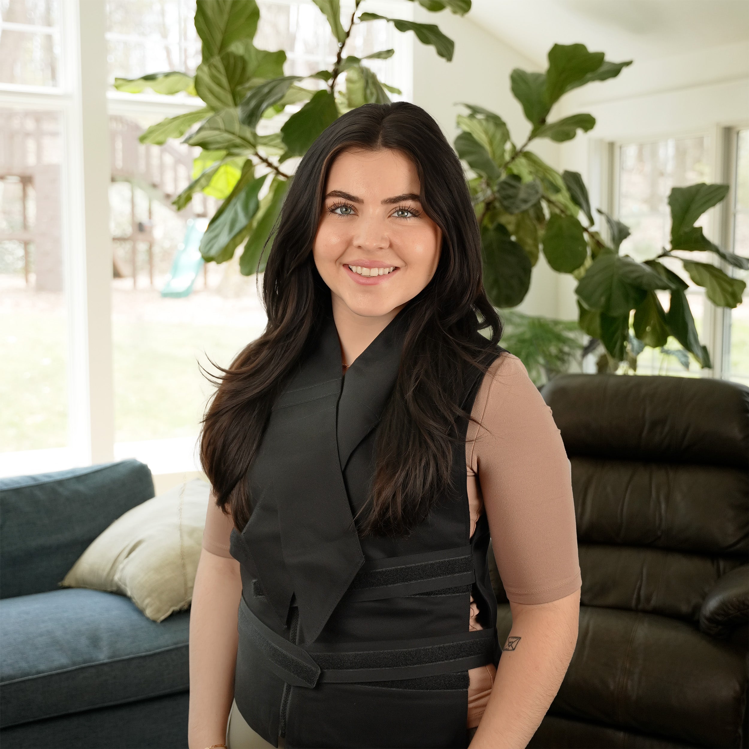 A woman with long dark hair stands indoors wearing a black Adjustable Zipper cooling vest and a Deluxe cooling neck wrap  over a sheer taupe short-sleeve top. She is posed in a bright living room with large green plants, a blue sofa, and a dark leather chair in the background.