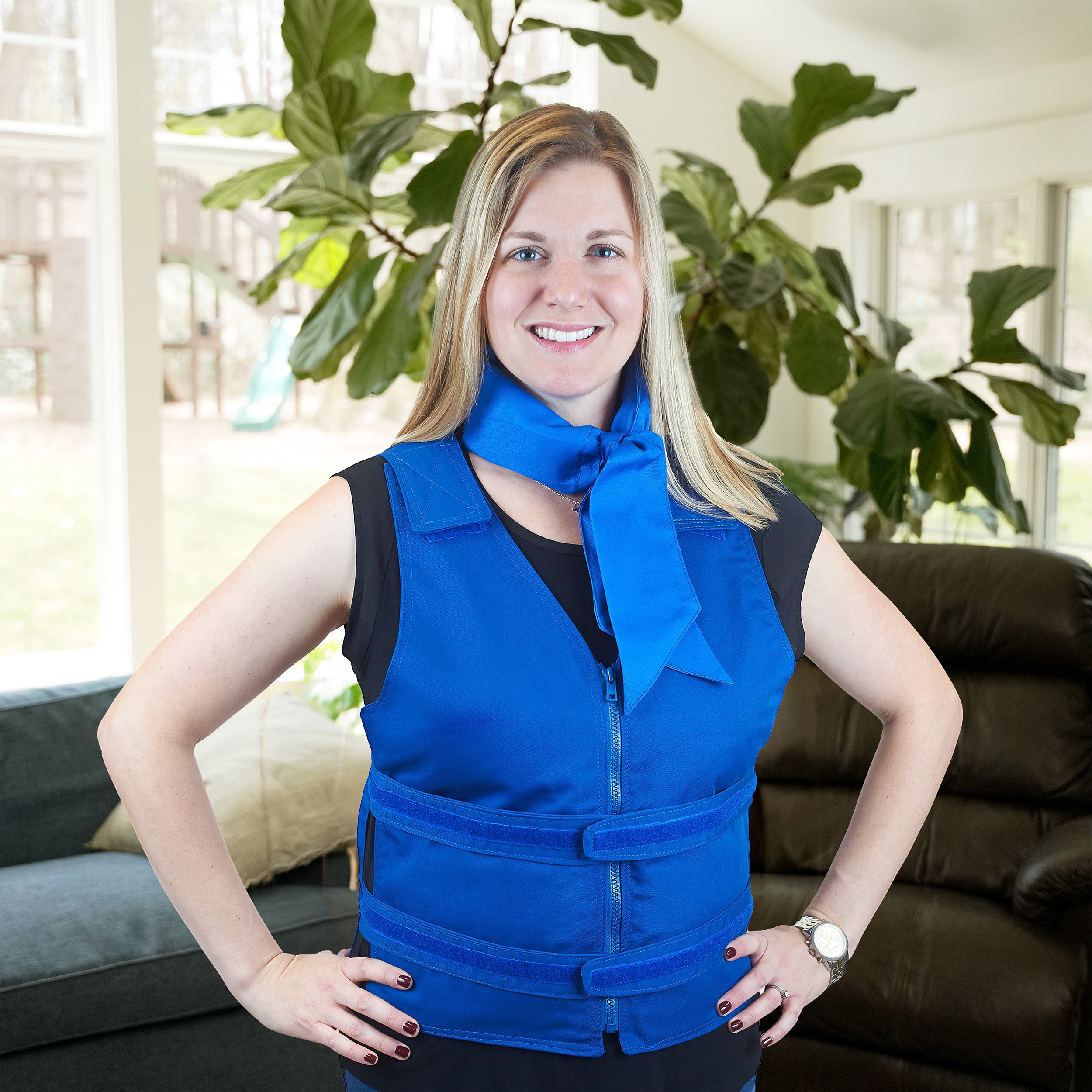 A smiling blonde woman stands indoors with her hands on her hips, wearing a bright blue adjustable zipper cooling vest and matching blue deluxe cooling neck wrap over a black top. The photo is taken in a living room with a couch, windows, and large green plants in the background.