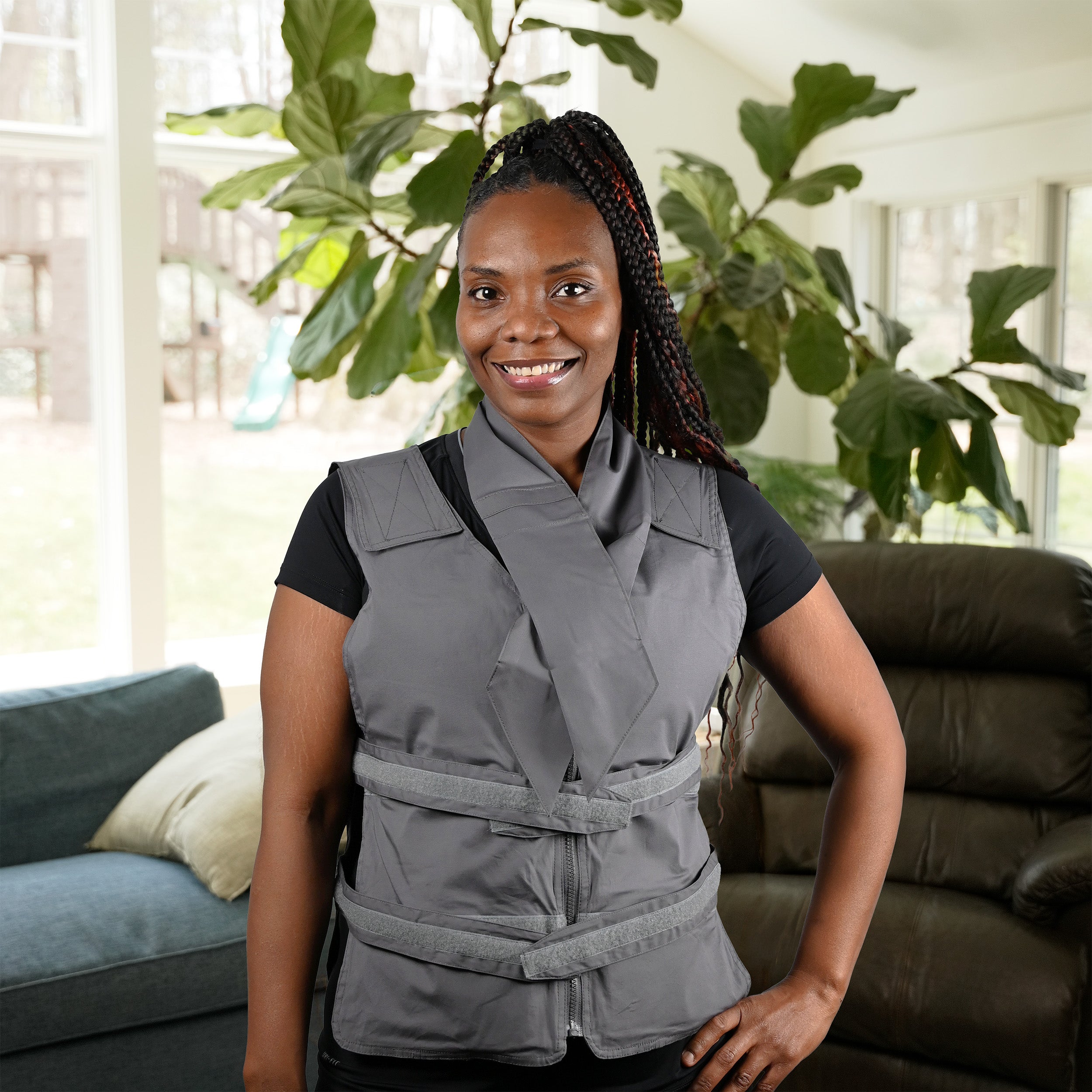 A smiling woman wearing a gray adjustable cooling vest over a black t-shirt, standing in a brightly lit living room with a large green plant in the background. She's also wearing a gray deluxe cooling neck wrap around her neck.