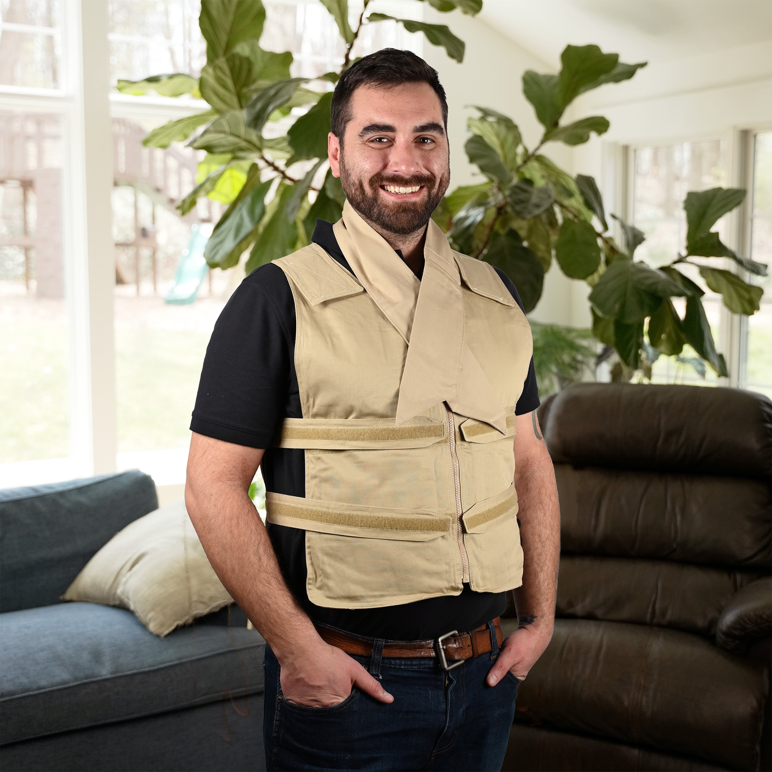 A smiling man wearing a beige adjustable cooling vest over a black t-shirt and jeans, standing in a living room with a large green plant in the background. He's also wearing a beige deluxe cooling neck wrap.