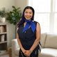 A woman with purple hair stands in a bright living room wearing an Adjustable zipper cooling vest in nylon with a blue deluxe cooling neck wrap and dark patterned bottoms.