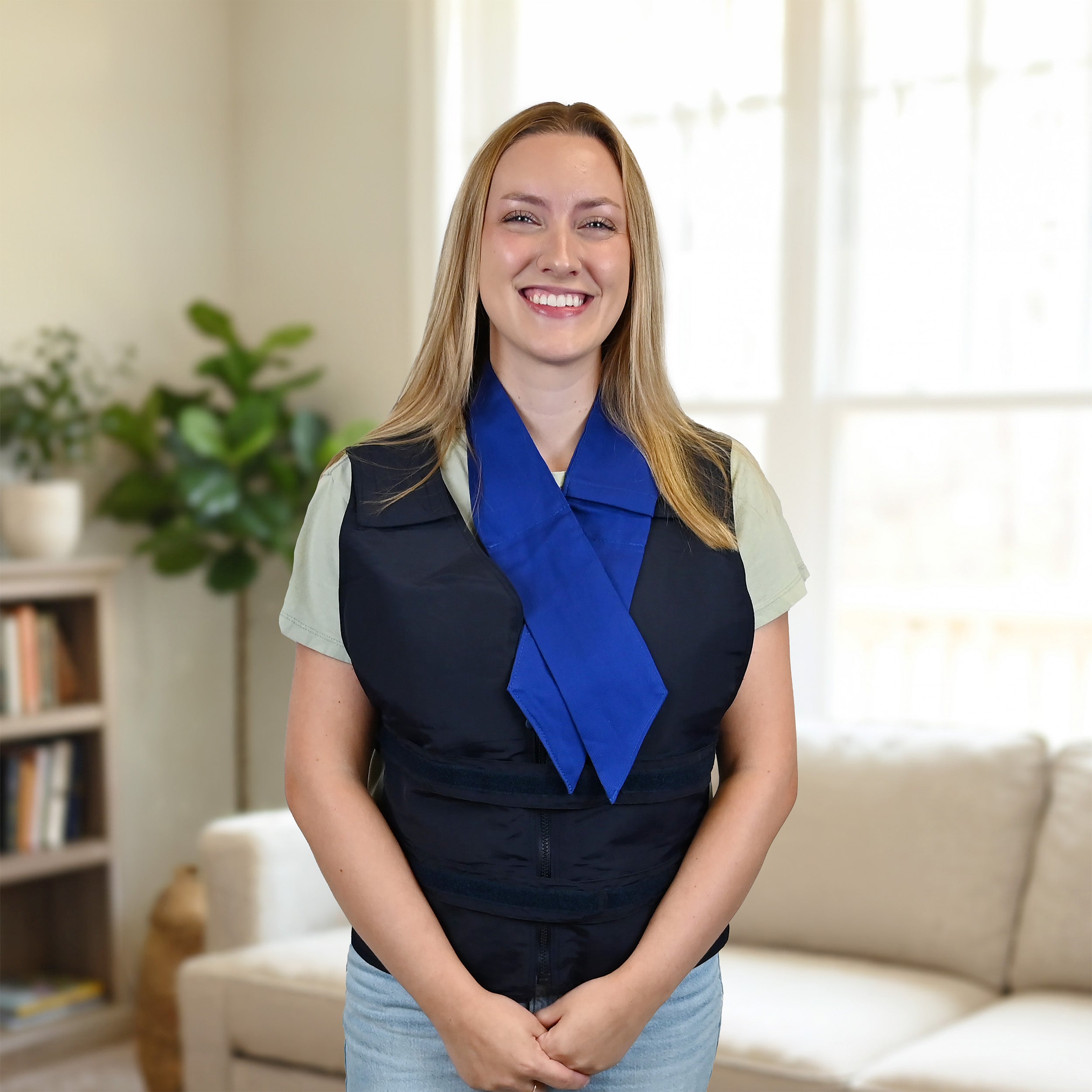 A smiling woman with blonde hair stands indoors wearing a dark navy adjustable cooling vest in nylon over a light sage green top and a blue deluxe cooling neck wrap. The background shows a softly lit living room with a sofa, bookshelf, and plant.