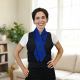 A smiling woman standing indoors with her hands on her hips, wearing a white short-sleeve top, a dark navy adjustable cooling vest, and a bright blue deluxe cooling neck scarf. The background shows a softly blurred living room with a sofa, bookshelf, and plant.