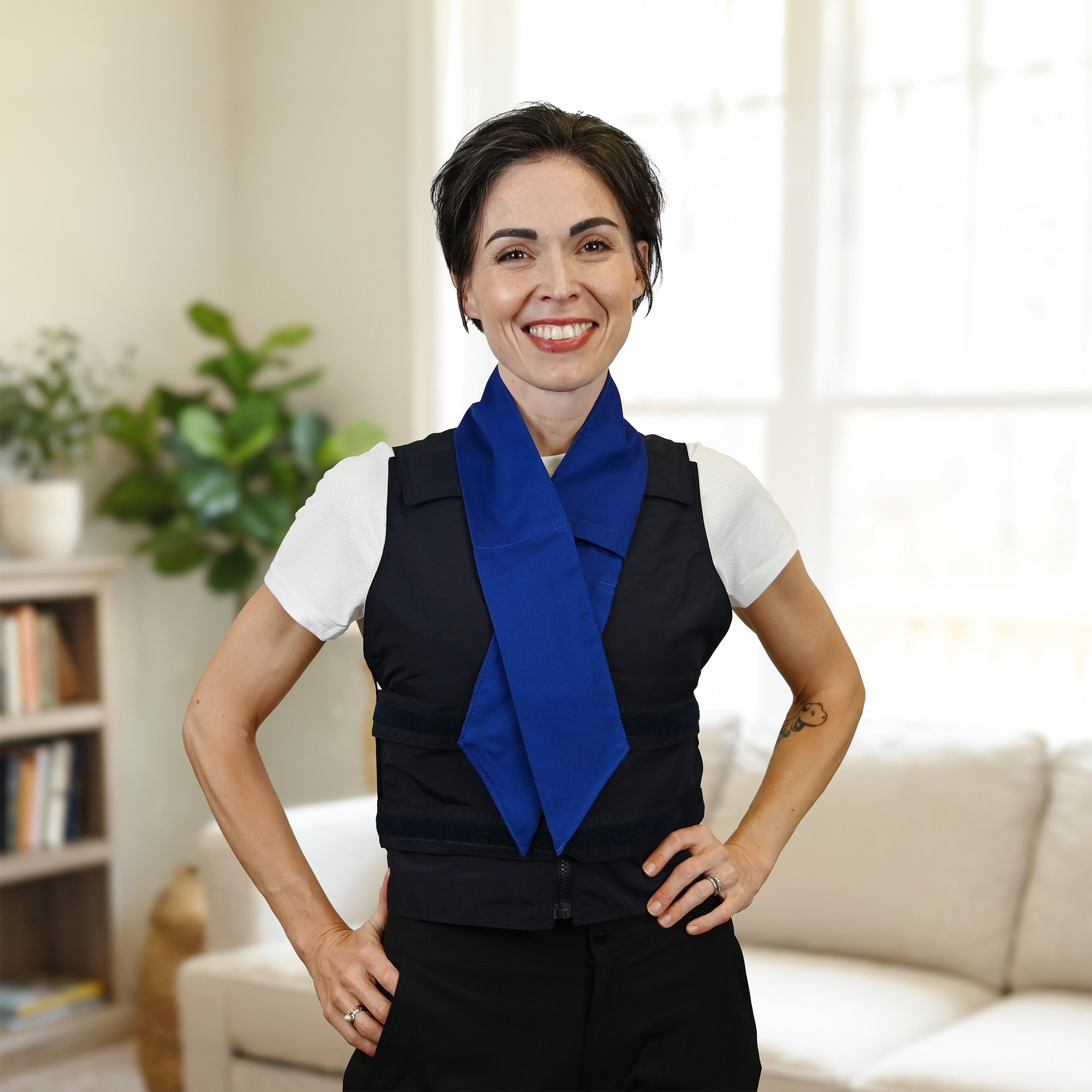 A smiling woman standing indoors with her hands on her hips, wearing a white short-sleeve top, a dark navy adjustable cooling vest, and a bright blue deluxe cooling neck scarf. The background shows a softly blurred living room with a sofa, bookshelf, and plant.