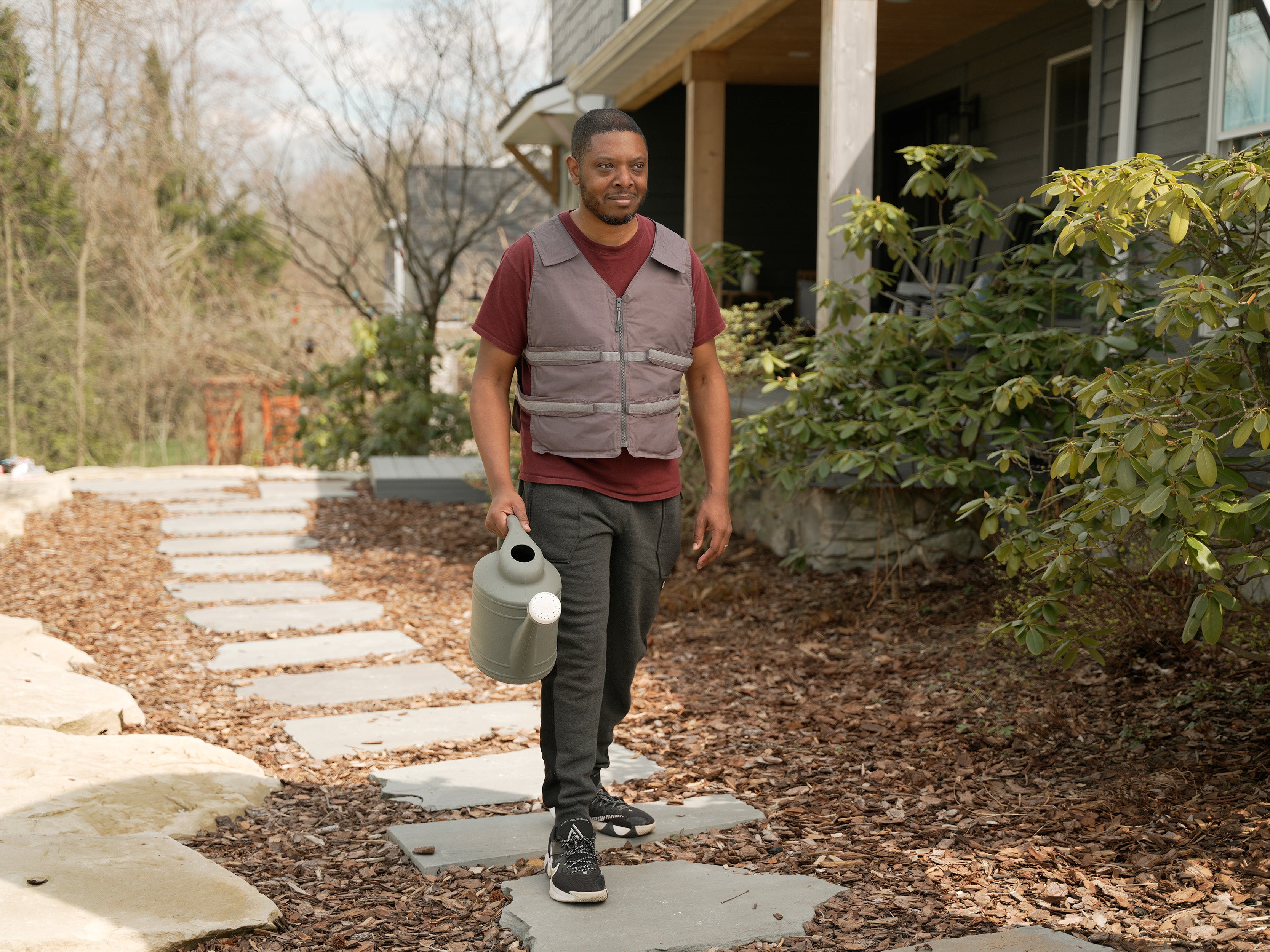 Man walking outdoors holding a watering can, with a house and garden in the background. He is wearing a gray adjustable cooling vest over a maroon t-shirt.