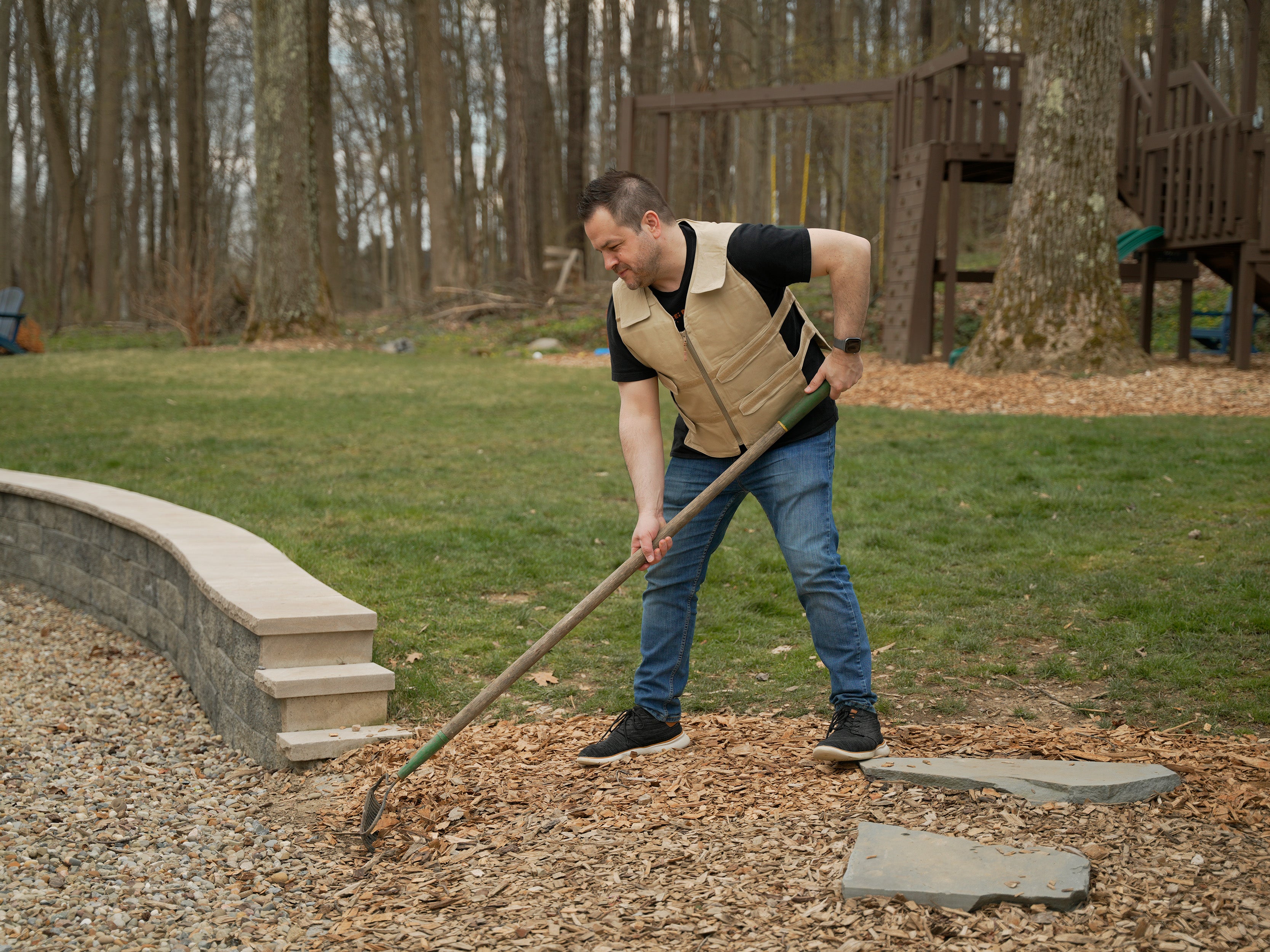 A man in a tan Adjustable Zipper Cooling Vest and blue jeans raking wood chips in a backyard with trees and a wooden playset in the background.