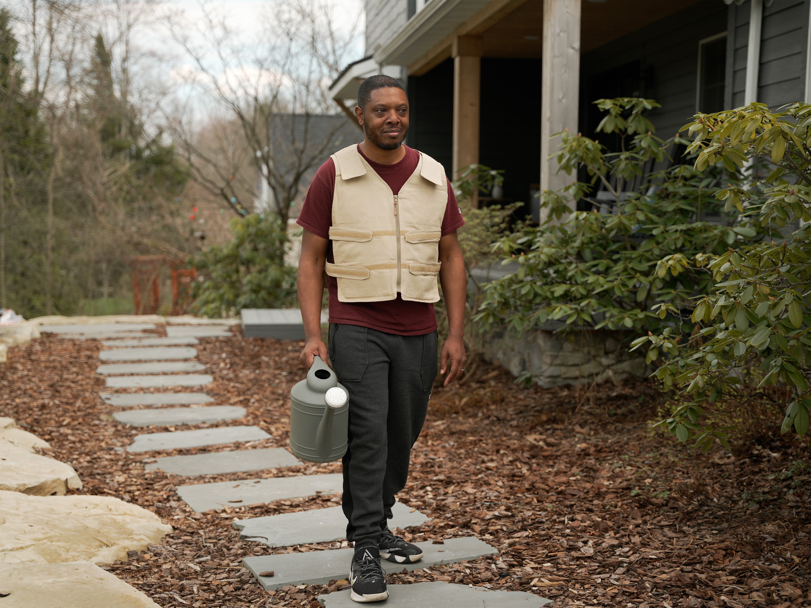 A man walks along a stone path in front of a house, wearing a beige adjustable cooling vest over a maroon t-shirt and dark pants, while holding a gray watering can.