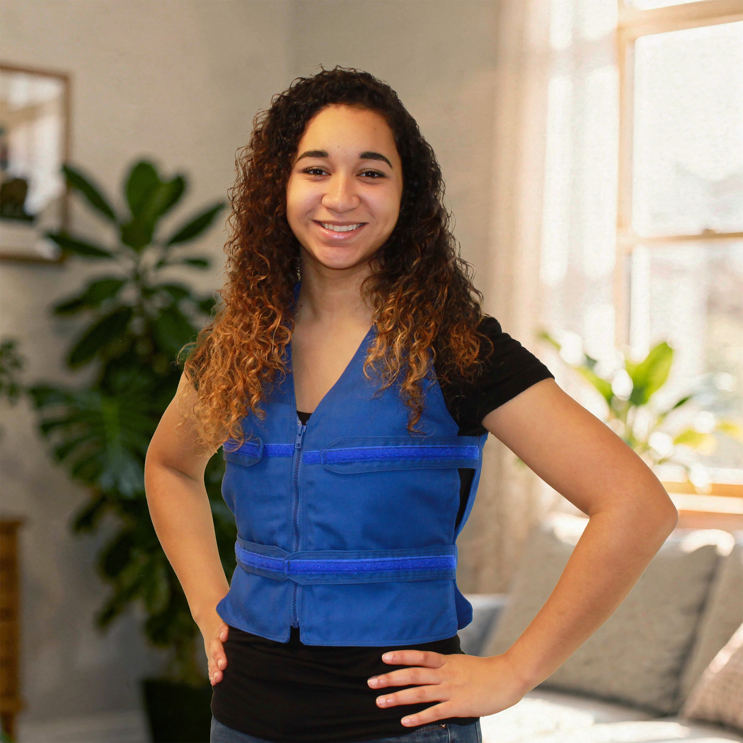 A smiling young woman with curly brown hair stands indoors wearing a bright blue adjustable cooling zip-front vest over a black short-sleeve top. The photo is taken in a softly lit room with plants and a window in the background.