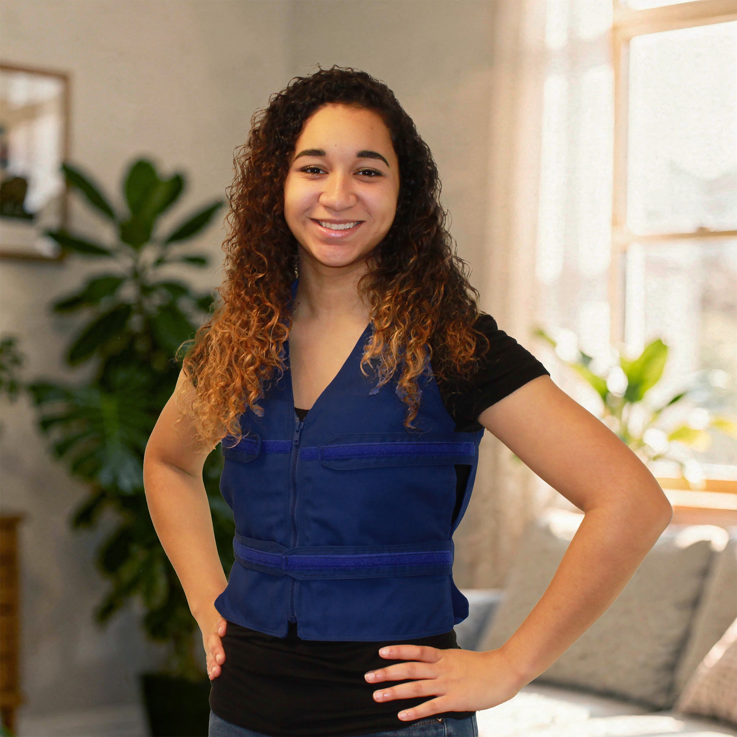 A smiling young woman with long curly brown hair stands indoors wearing a royal blue adjustable cooling zippered vest over a black short-sleeve top. The photo is taken in a softly lit room with plants and a window in the background.